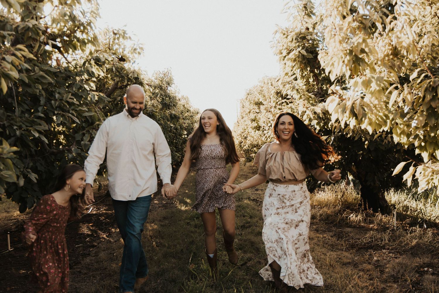 Family of four joyfully walking hand-in-hand through a lush orchard on a sunny day.
