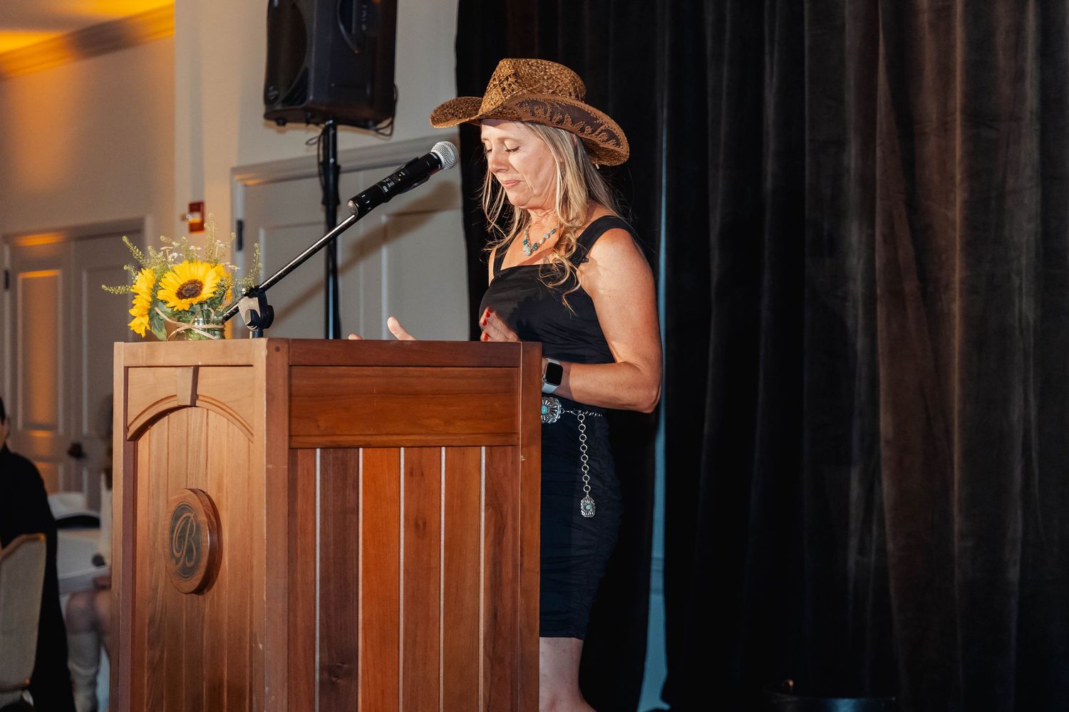 Woman in cowboy hat speaking at wooden podium with microphone at formal indoor event with sunflowers.