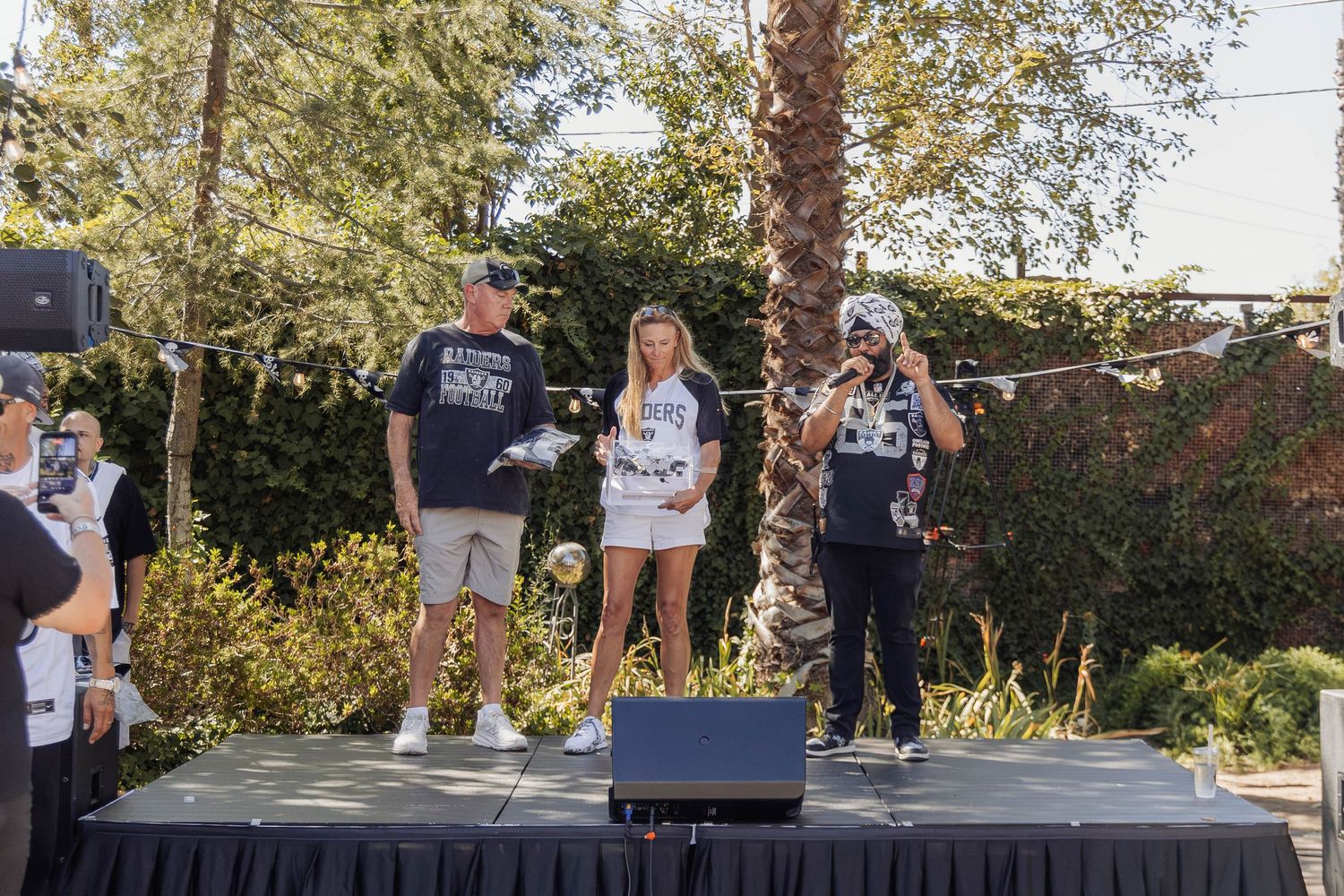 Three people stand on an outdoor stage at a casual event, surrounded by lush greenery on a sunny day.