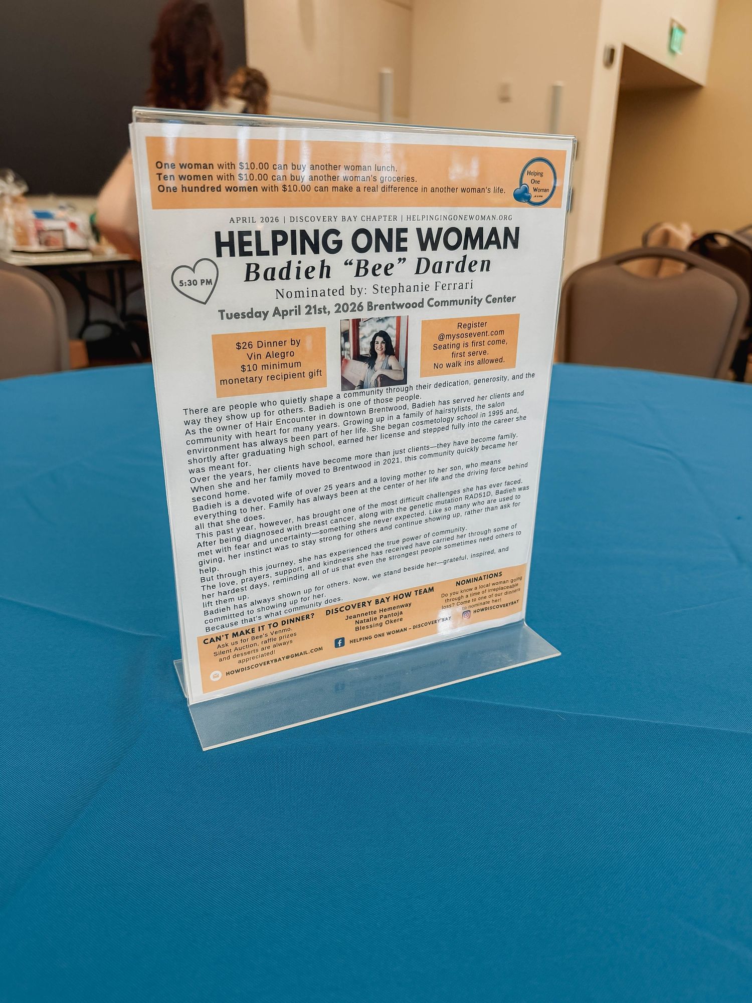 Acrylic sign on blue tablecloth promoting Helping One Woman event honoring Badieh Bee Darden on April 21st 2024.
