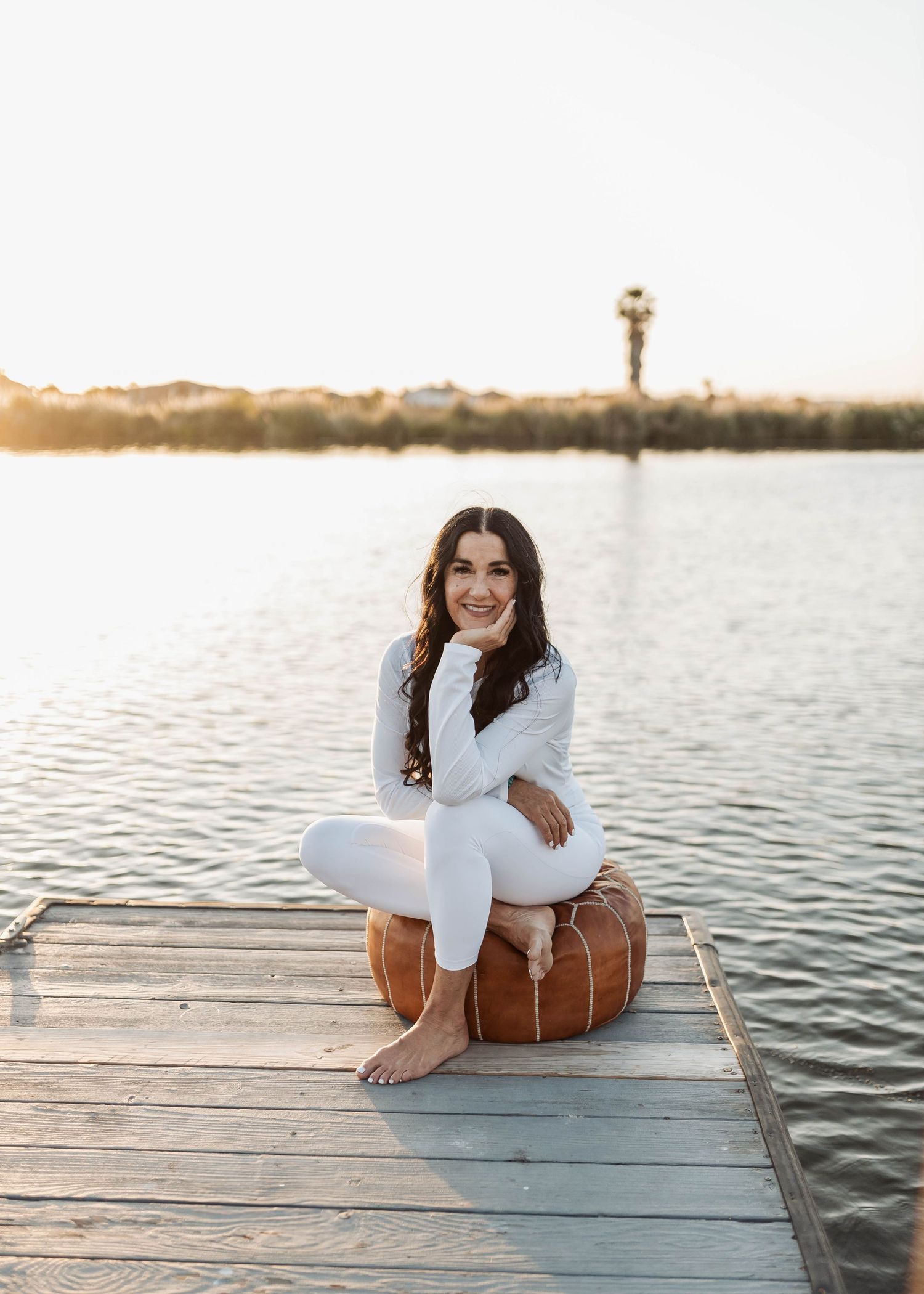 Woman sitting cross-legged on a brown leather ottoman on a waterfront dock.