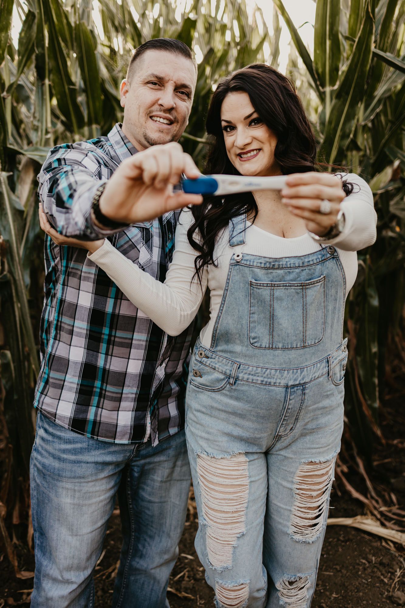 Couple excitedly holding positive pregnancy test in front of cornfield backdrop.