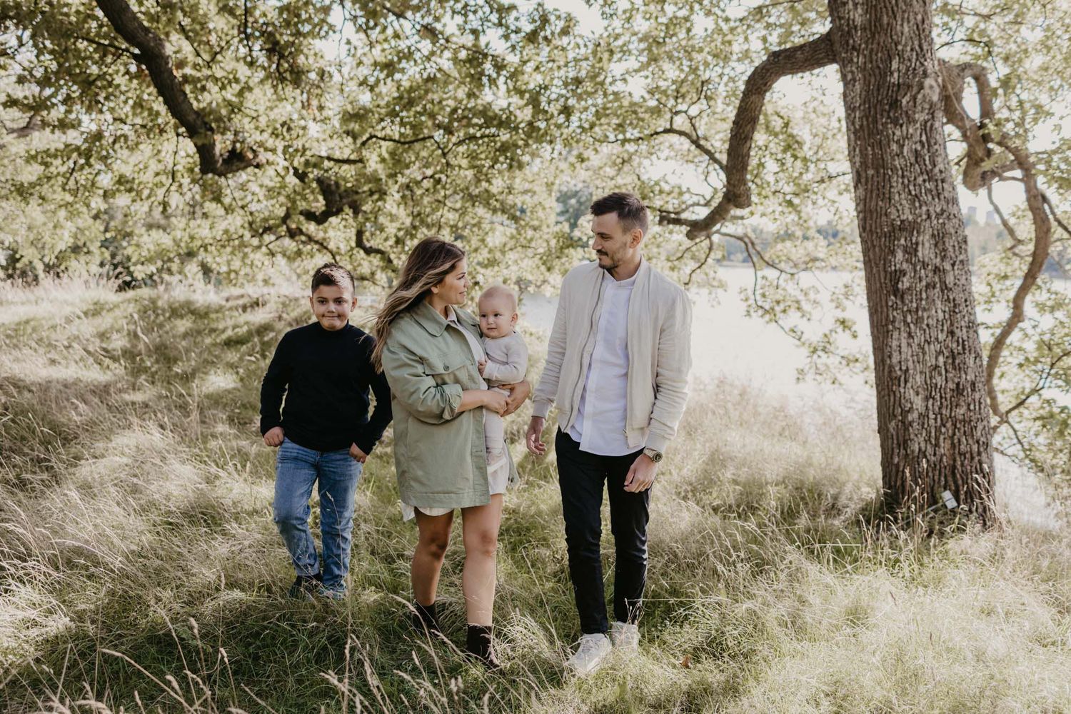 A casual outdoor family photoshoot in a sunlit grassy area under oak trees.