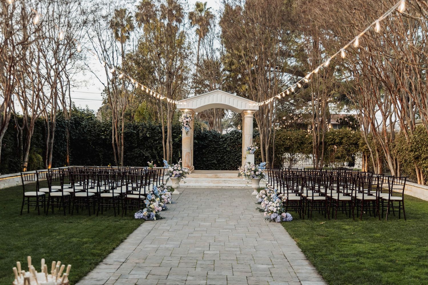 Elegant outdoor wedding ceremony setup with white arch, string lights, black chairs, and floral arrangements on a manicured lawn.