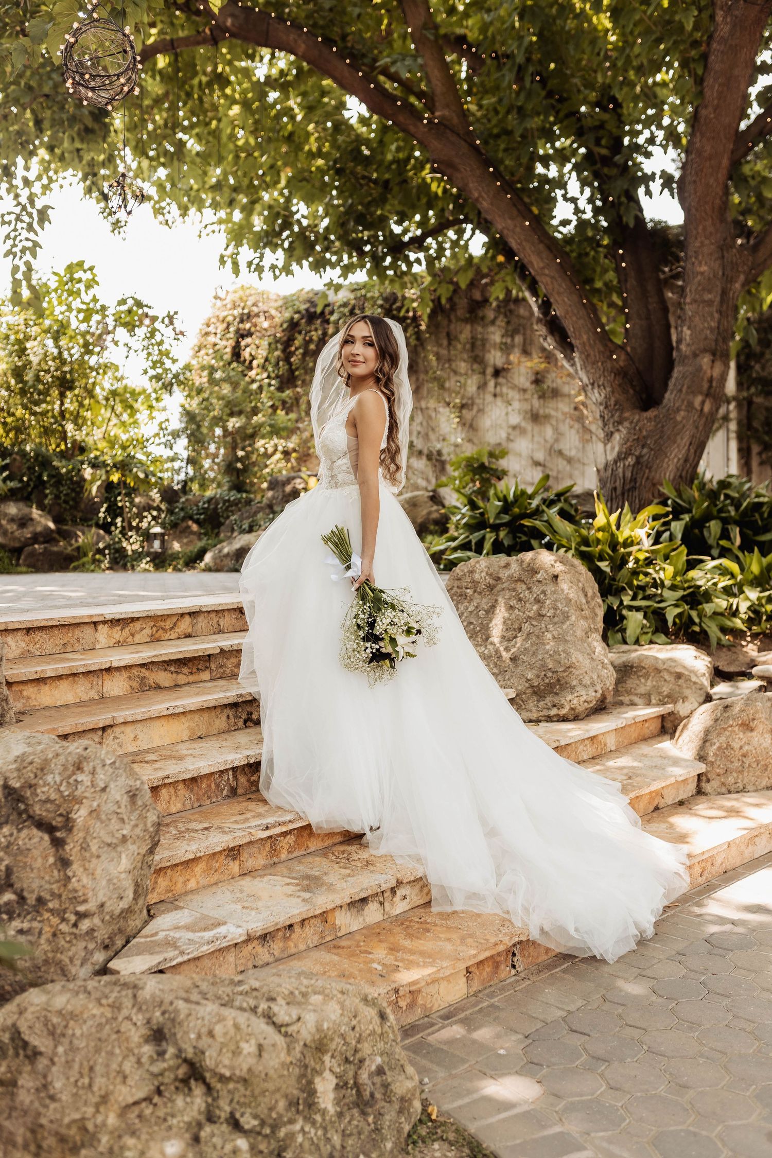 An elegant bride in a voluminous white dress poses on rustic wooden steps surrounded by lush greenery.