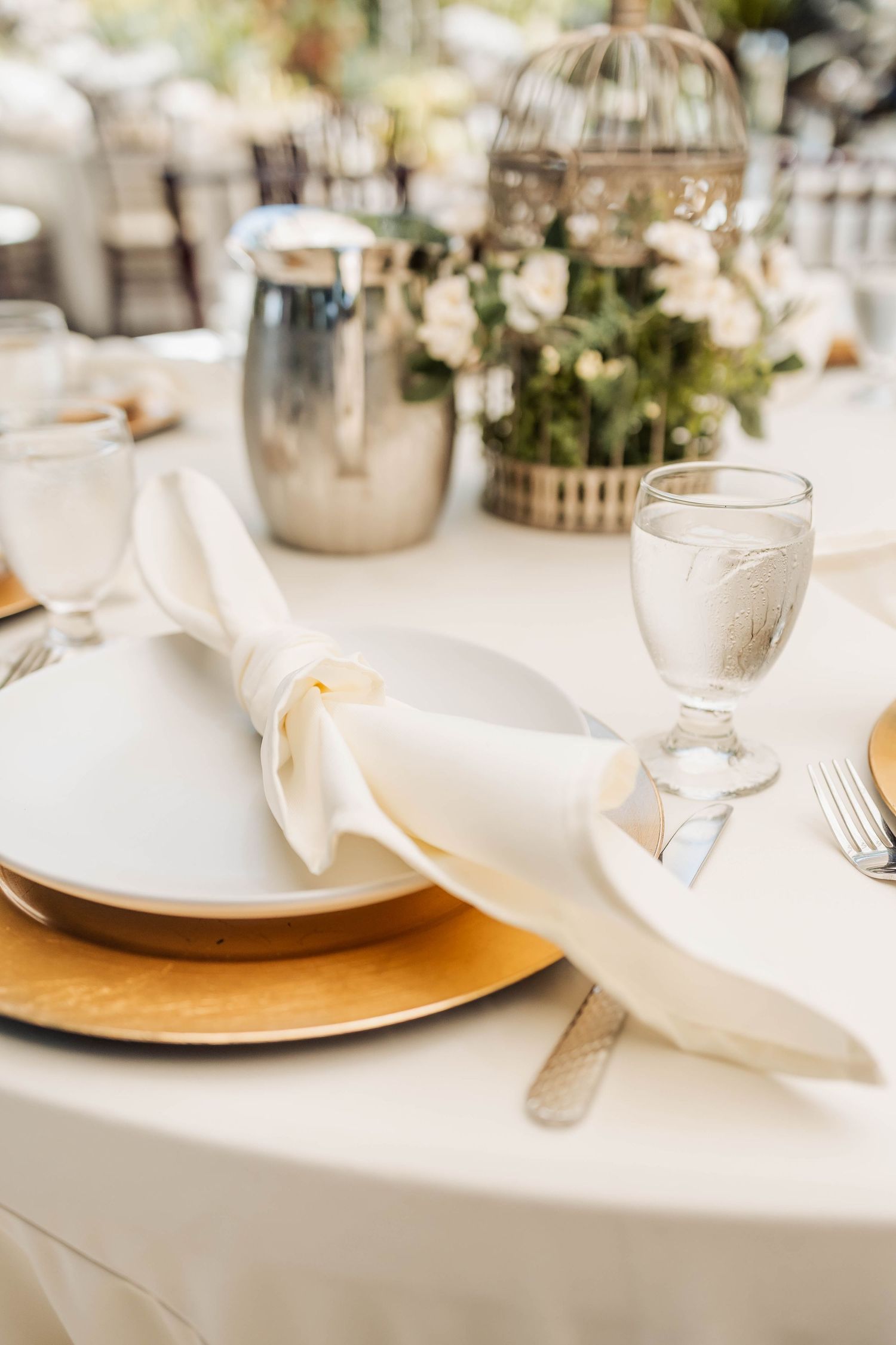 Elegant table setting with gold charger plates, white linens, and soft focus floral centerpieces for a wedding reception.