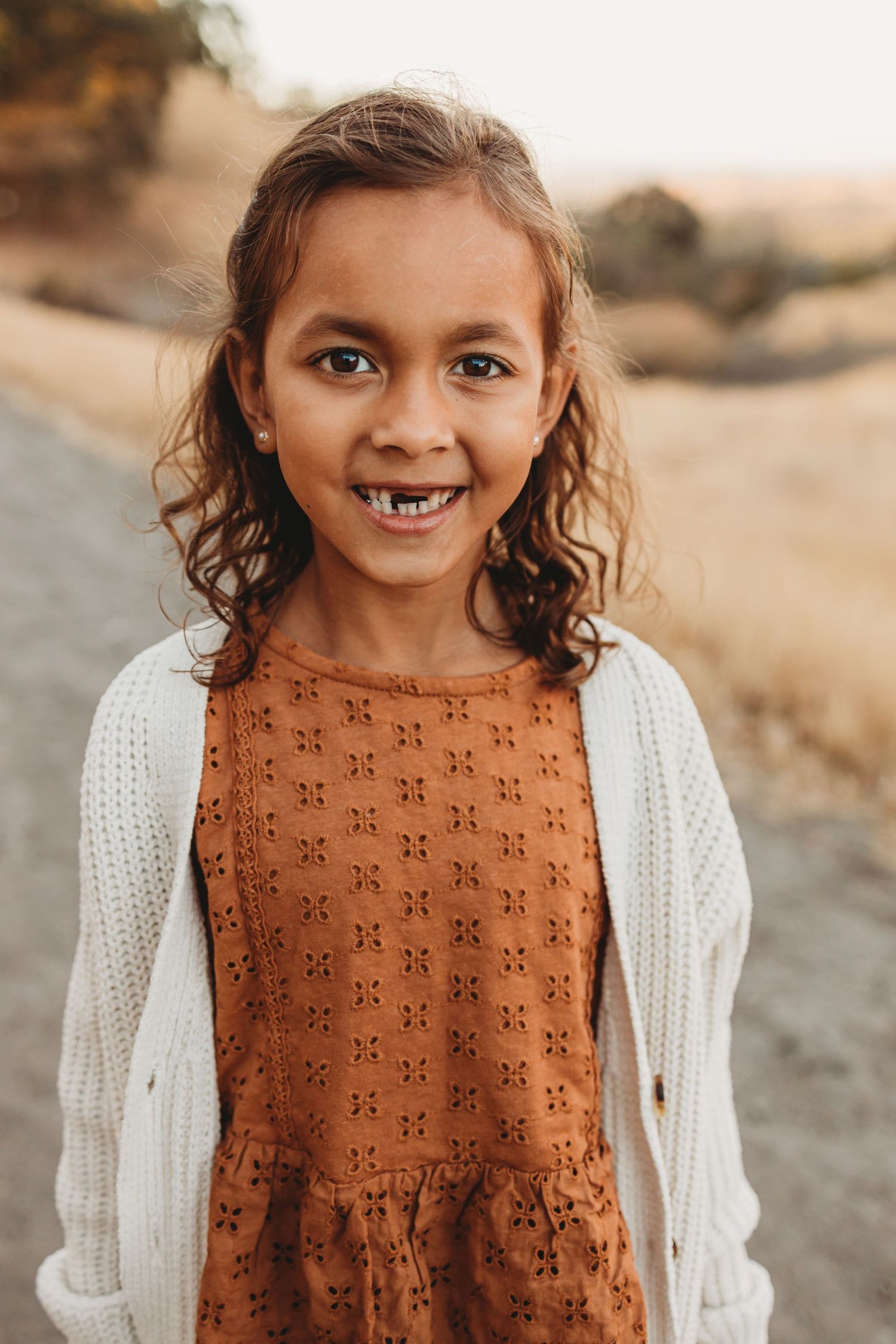Young person wearing white cardigan and brown patterned dress smiling at camera on nature trail.