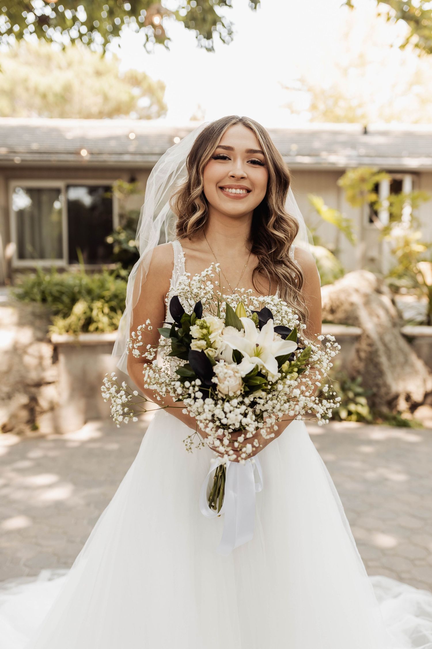 A bride in a white lace wedding gown holds a stunning bouquet of white lilies and baby's breath in a garden setting.