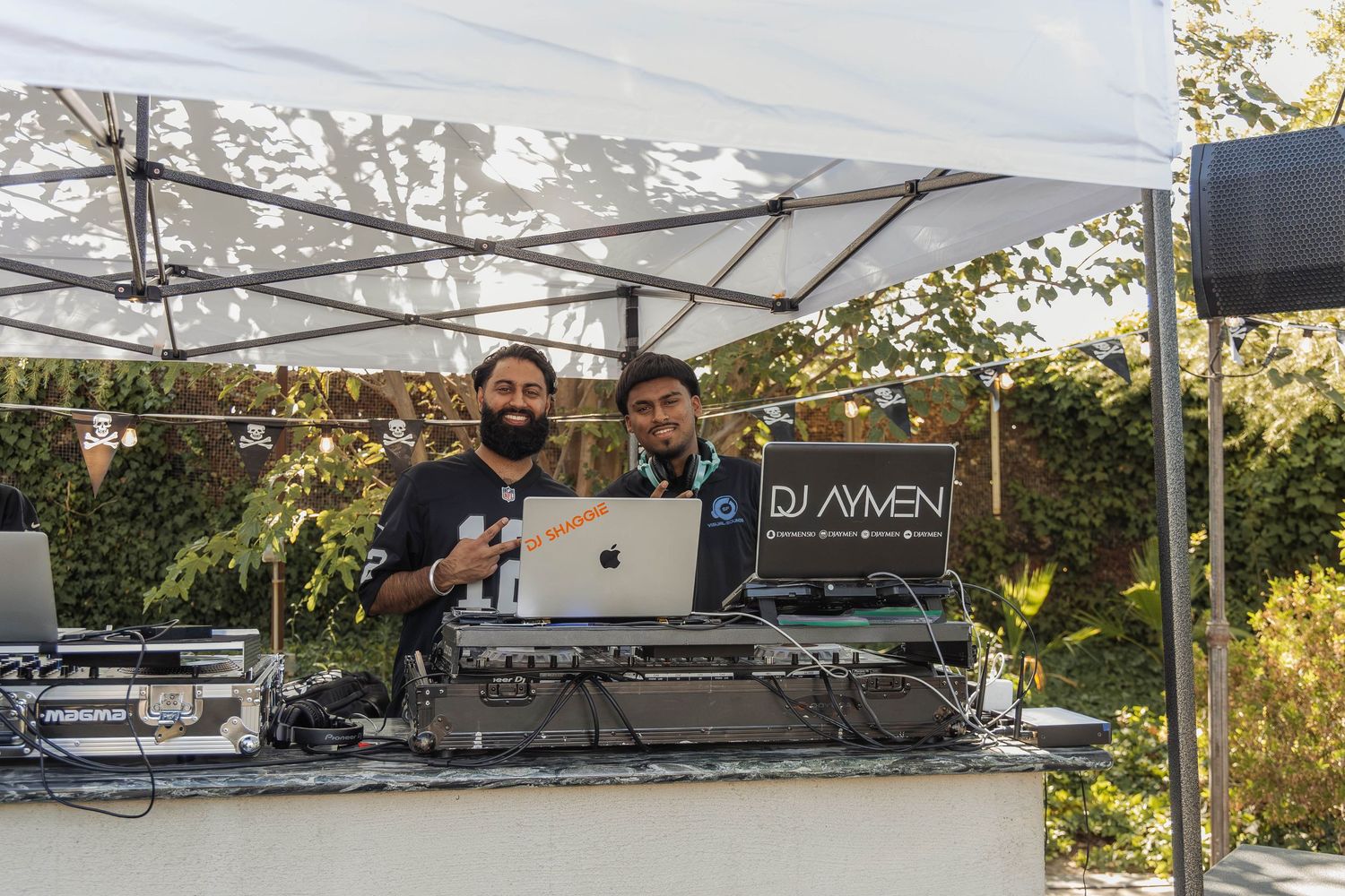 Two DJs posing behind turntables and laptop at DJ Aymen setup under white canopy outdoors.