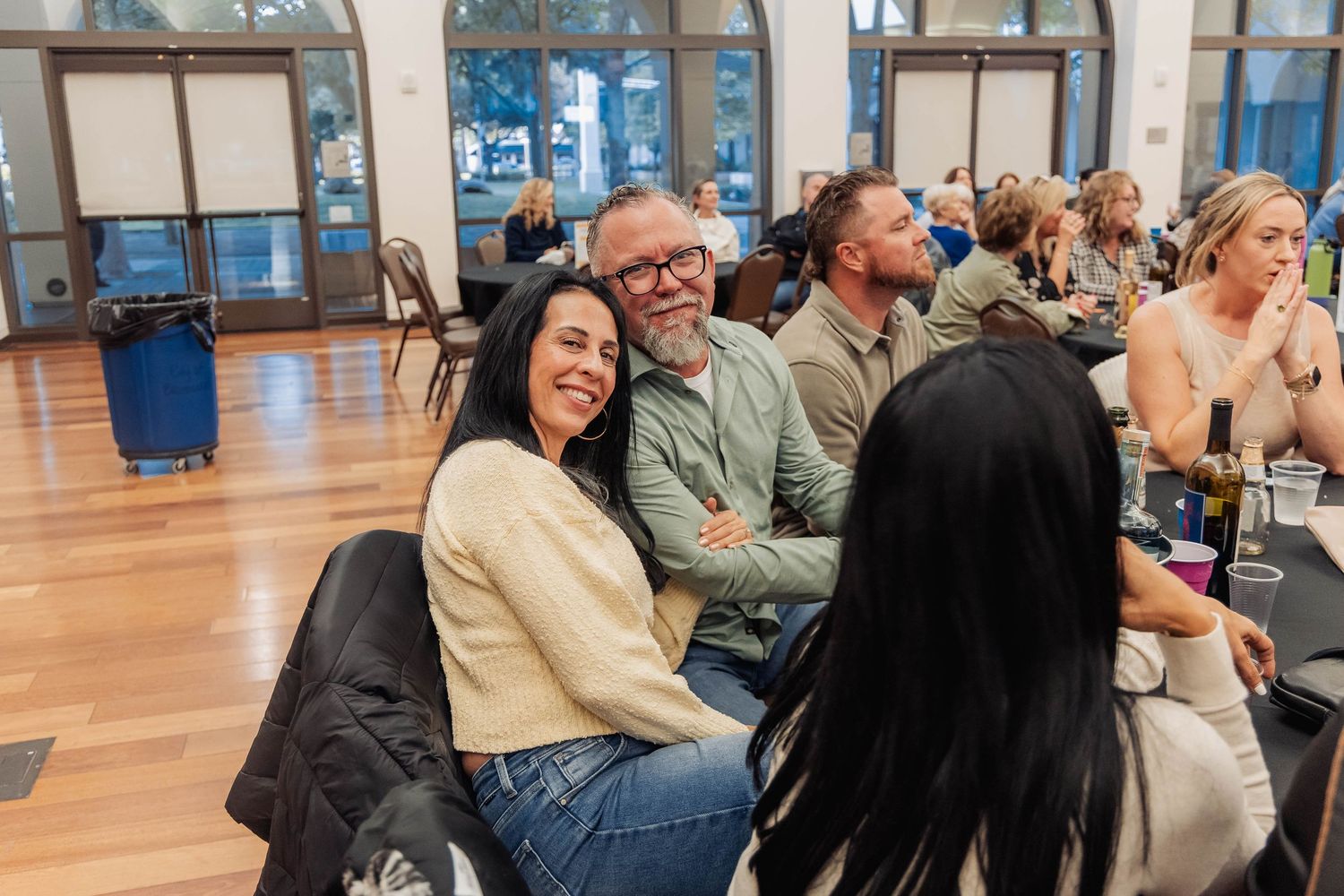 People smiling and socializing at an indoor event in a bright hall with tables and guests in the background.