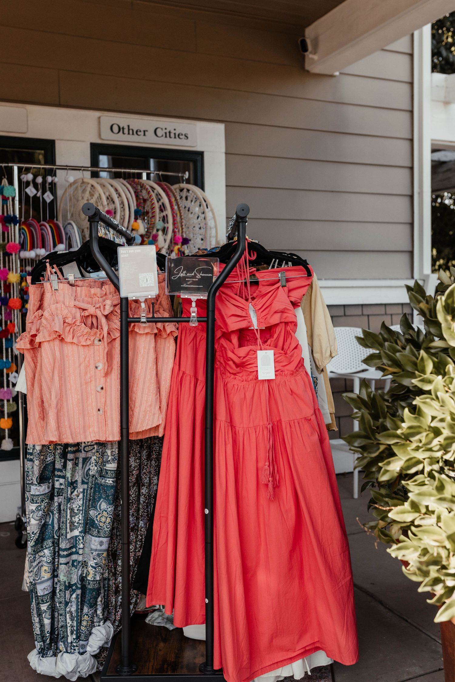 Clothing rack displaying pink and coral summer dresses and casual wear outside a boutique named 'Other Cities'.