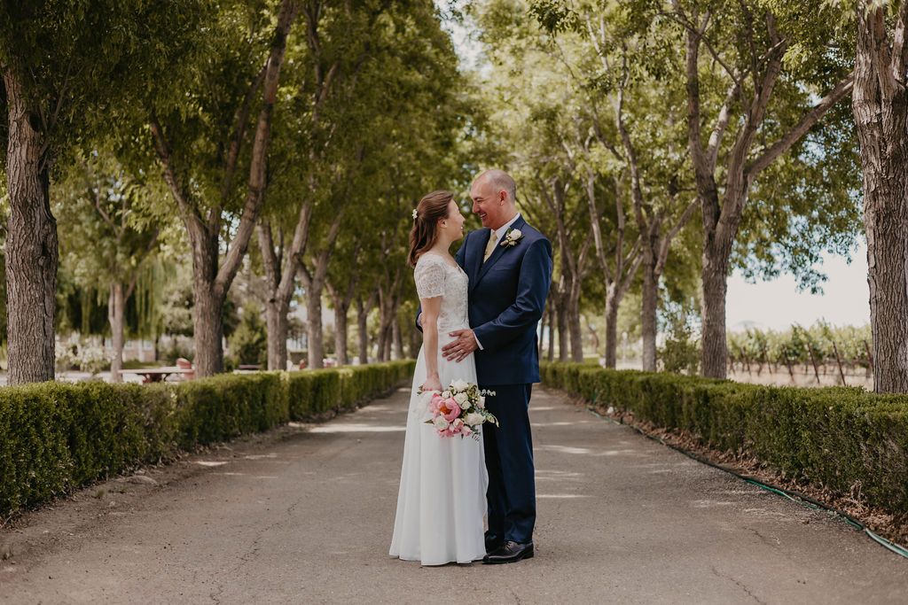 Newlyweds share an intimate moment on a tree-lined path in a garden wedding venue with manicured hedges.