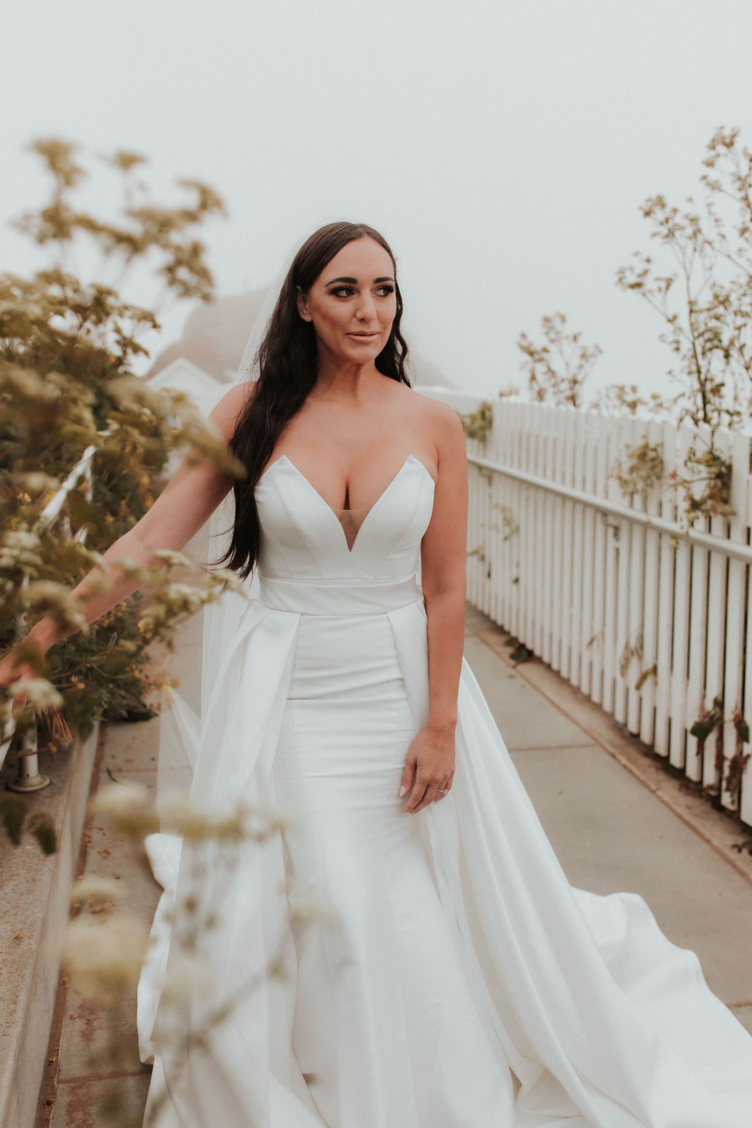 Bride in fitted wedding dress poses on pathway with white picket fence.