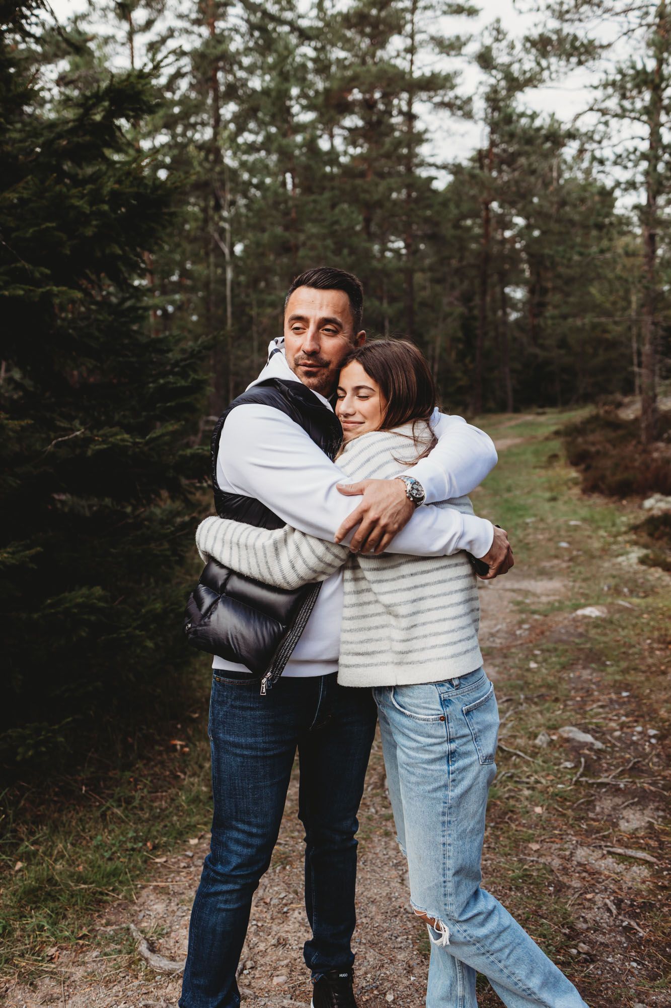 Pair embracing warmly while standing on a nature trail surrounded by pine trees.