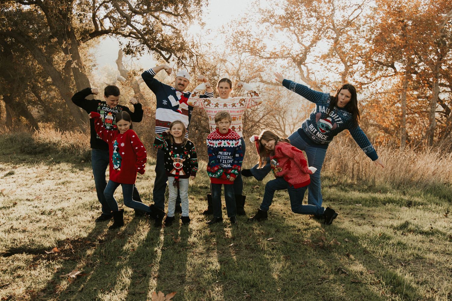 Group of children in Christmas sweaters posing playfully outdoors in a field with autumn trees.