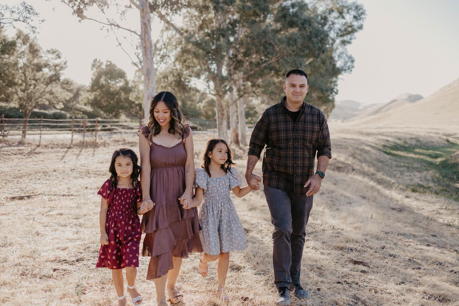 Family of four walking hand-in-hand on a sunny dirt path surrounded by trees and hills.