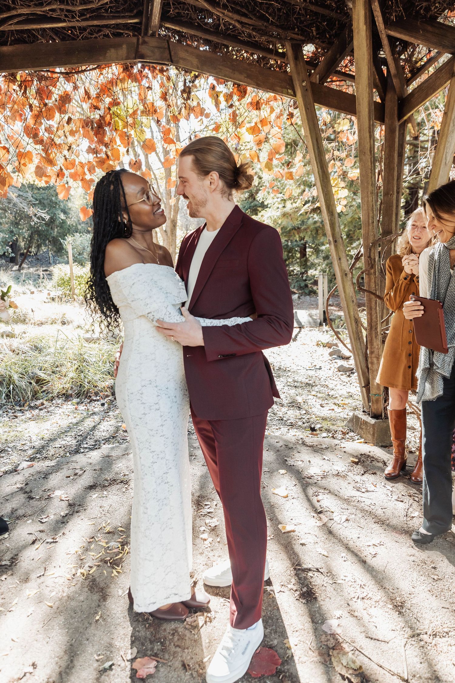 Dancing couple in white lace and burgundy suit under rustic wooden pavilion.