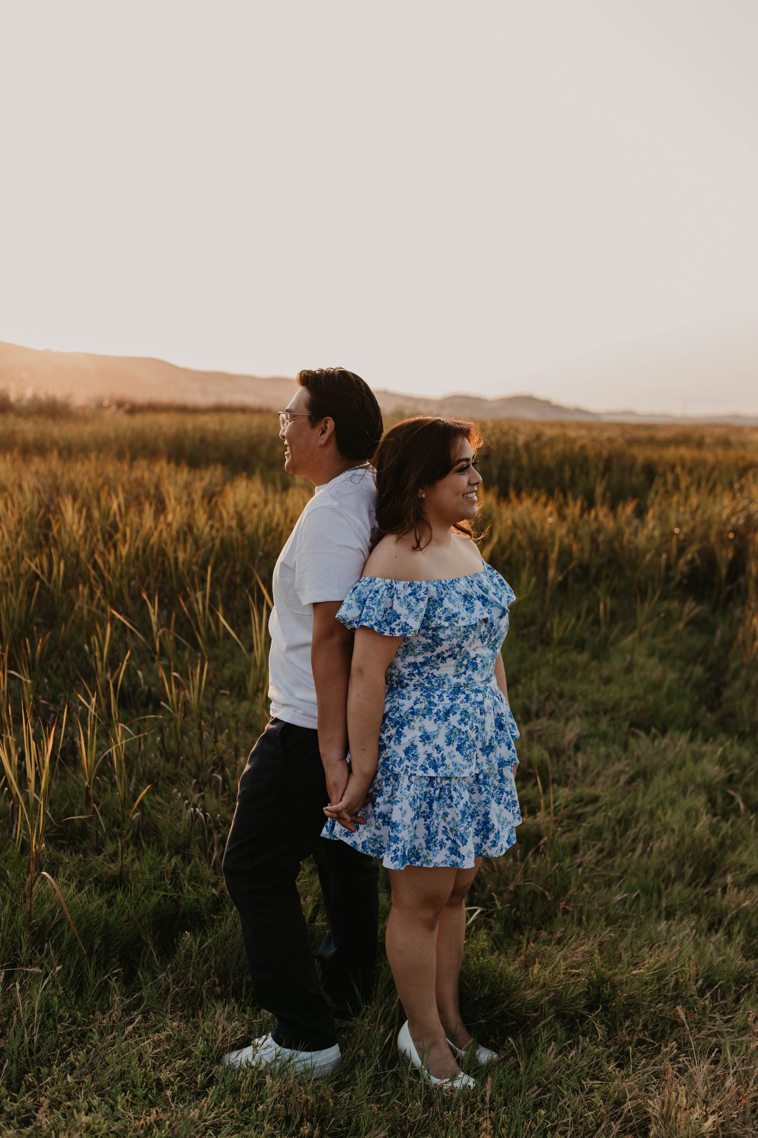 Couple in white and blue floral outfits walking through sunset-lit field.
