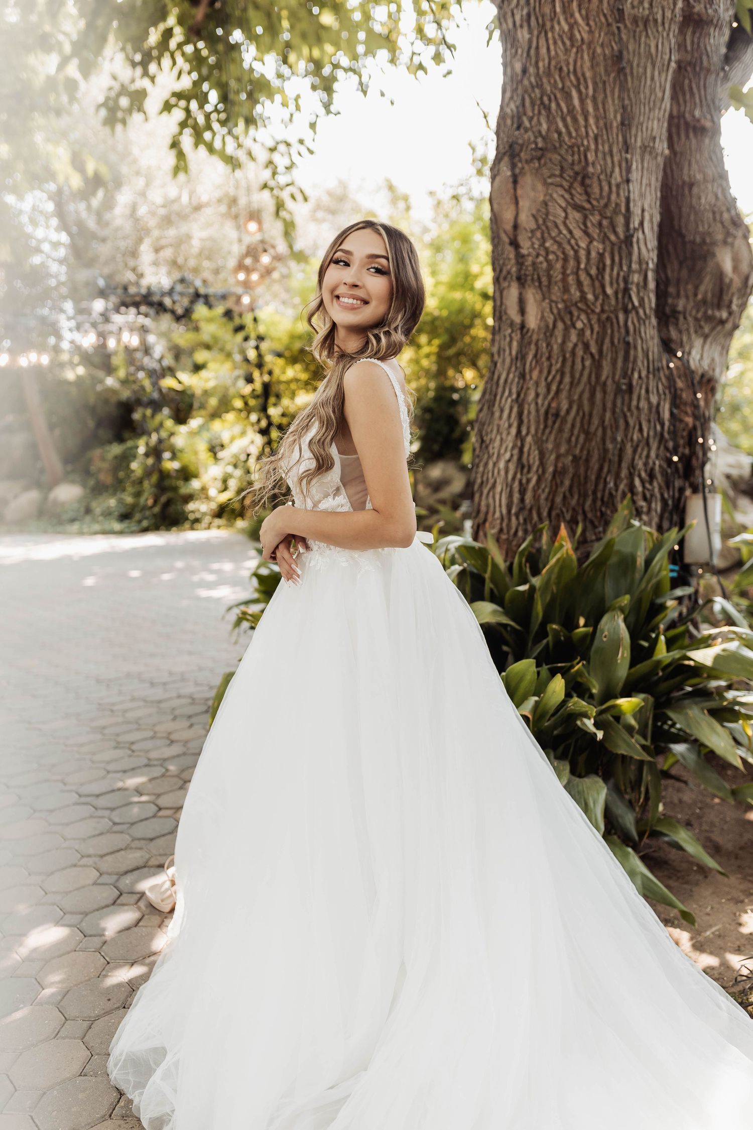 A bride in a voluminous white gown poses near a large tree trunk in a garden setting.