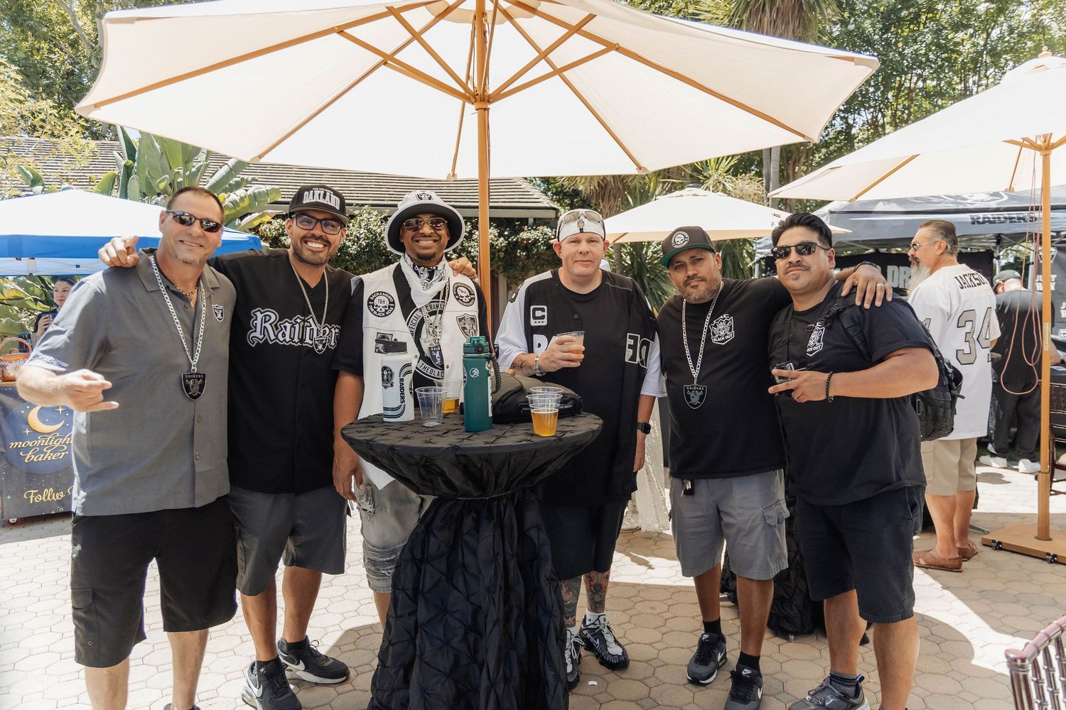Group of Raiders fans posing together outdoors under a white patio umbrella at a sunny tailgate party event.