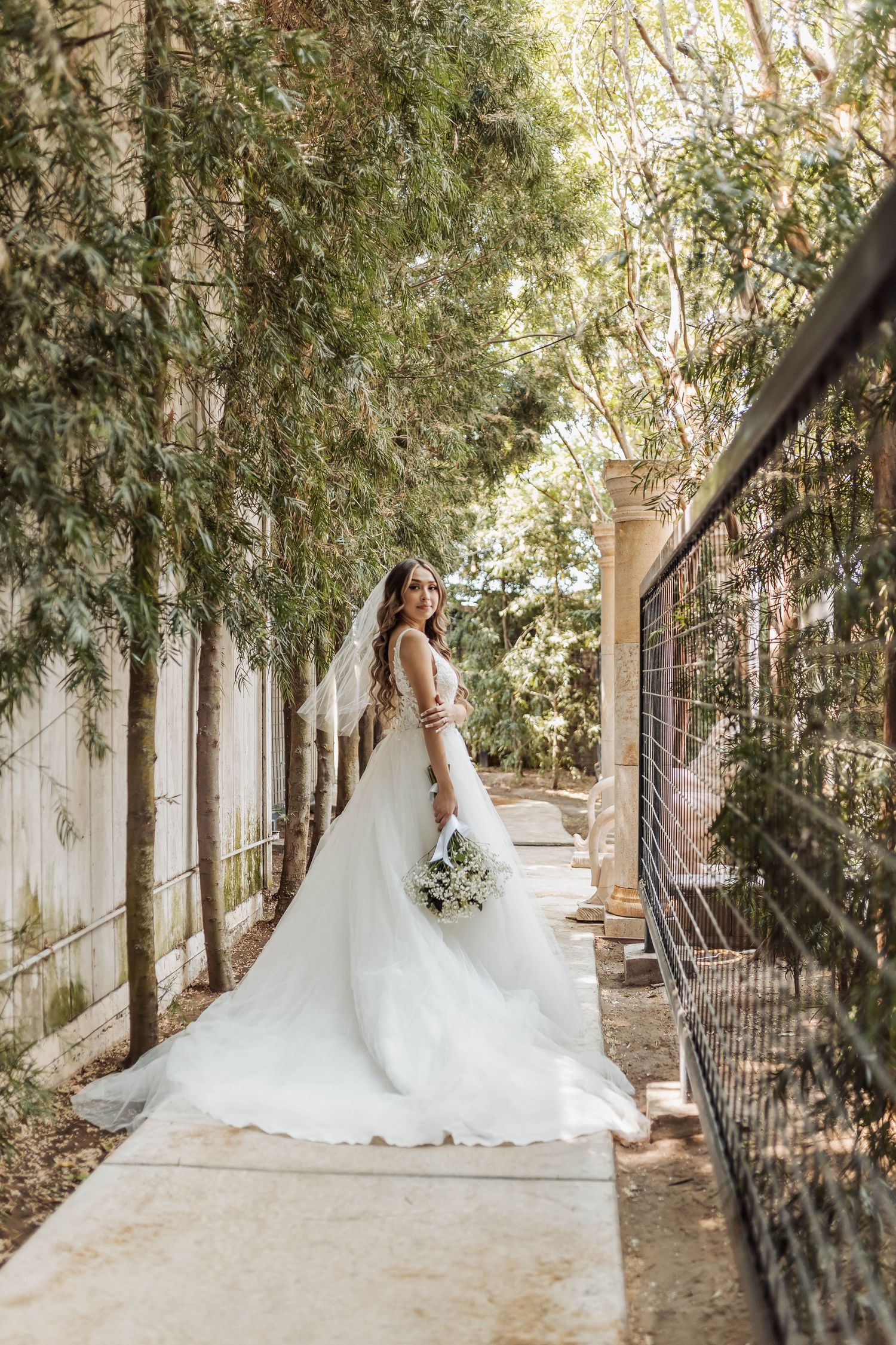 A beautiful wedding dress silhouette captured along a tree-lined pathway with white pillars.