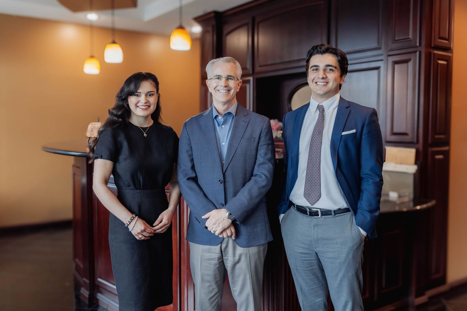 Three people in business attire standing together in an office setting with wood paneled walls.