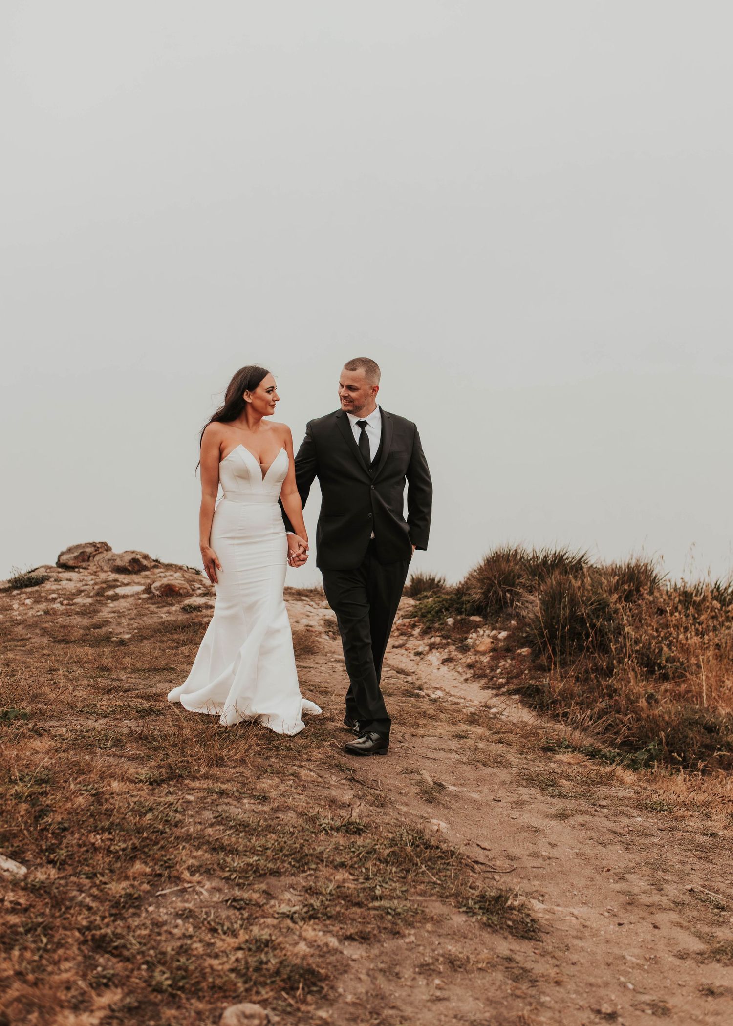 A couple walks hand in hand along a scenic coastal trail during their wedding day.
