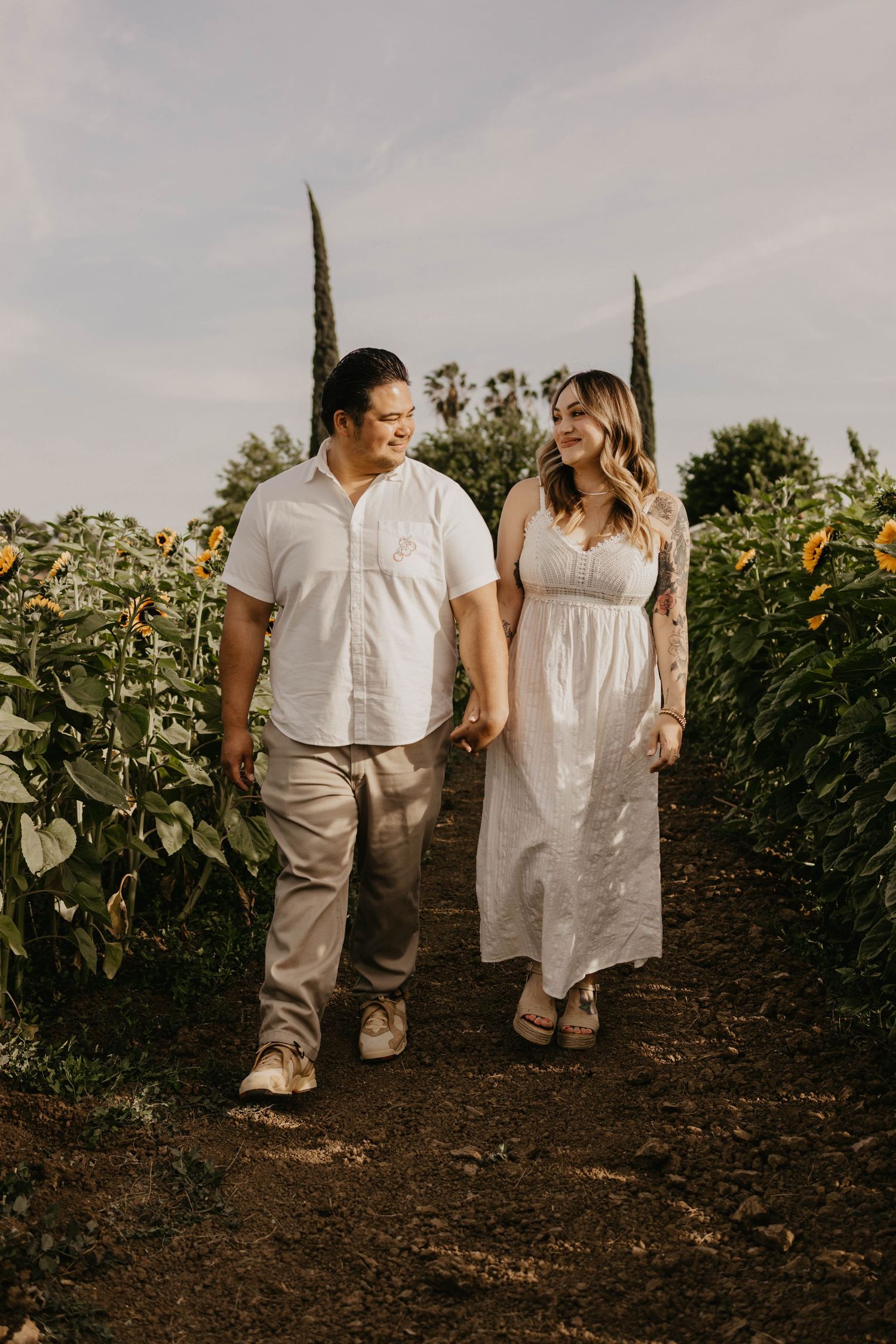 Couple poses intimately among rows of tall sunflowers at sunset wearing matching white and neutral outfits.