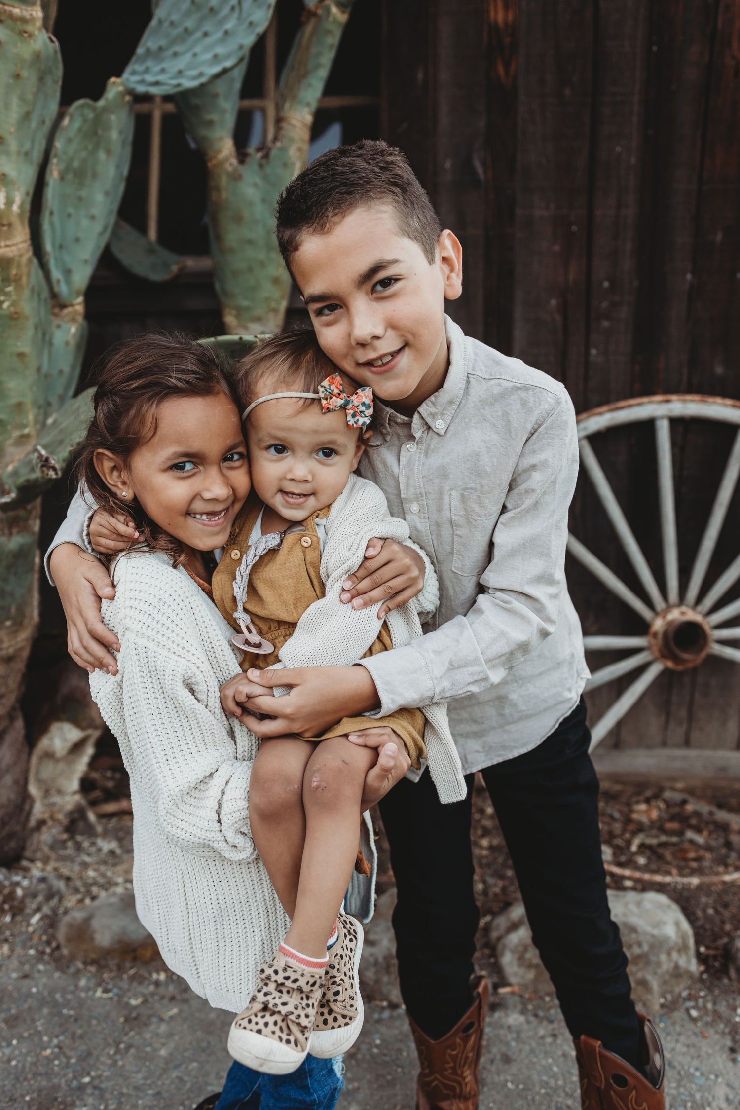Three children embracing and smiling together near wooden building and cactus.