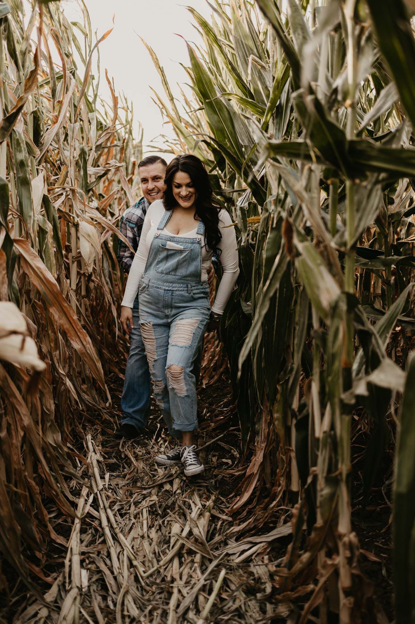 A couple poses together on a rustic path through tall autumn corn stalks.