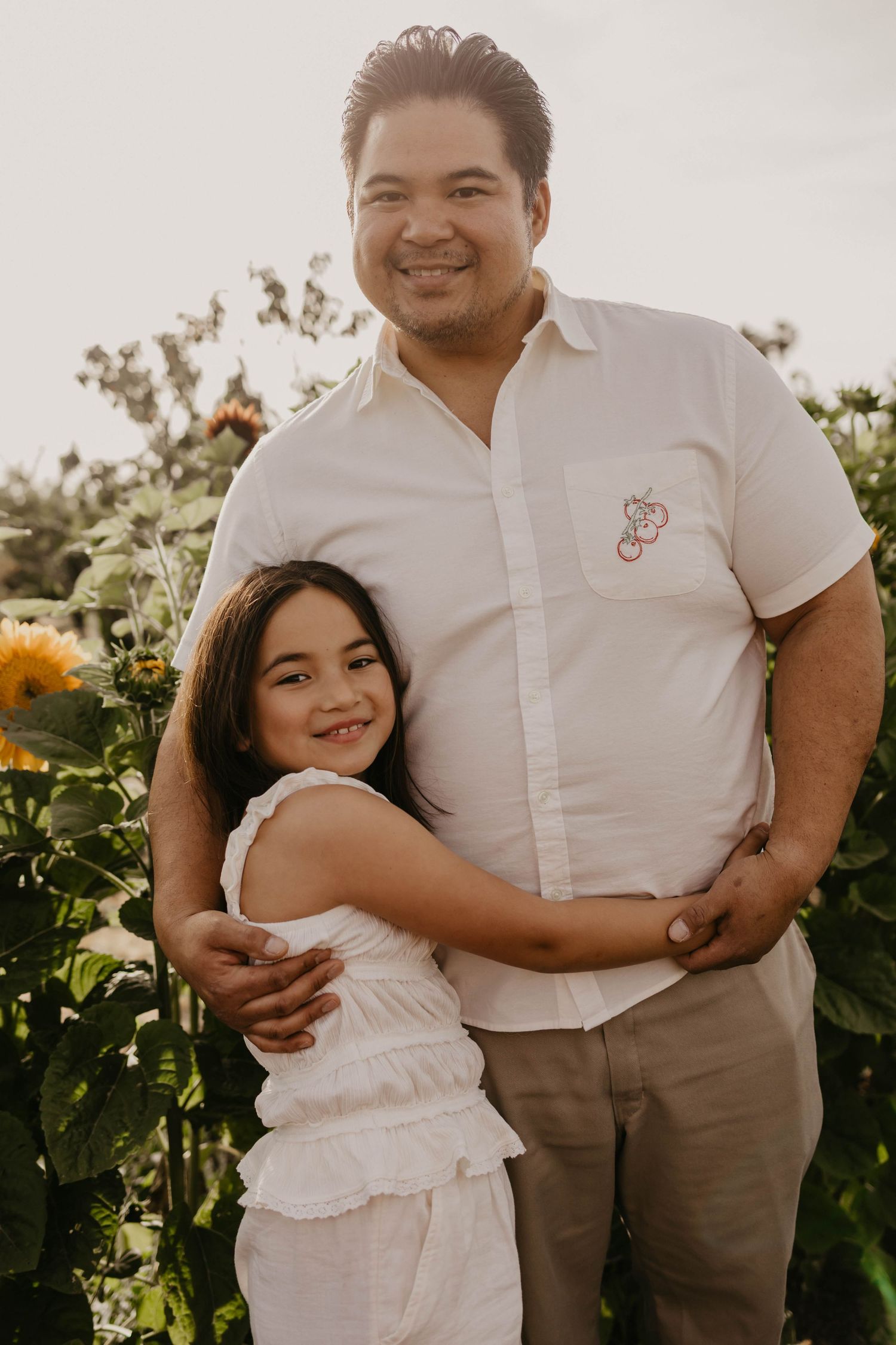 Family embraces warmly in sunflower field during golden hour wearing coordinated white and khaki outfits.