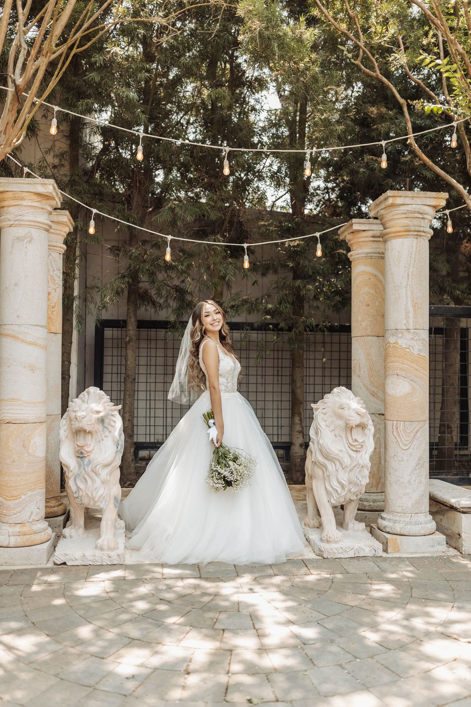 Full-length bridal portrait between stone pillars with lion statues and hanging string lights.