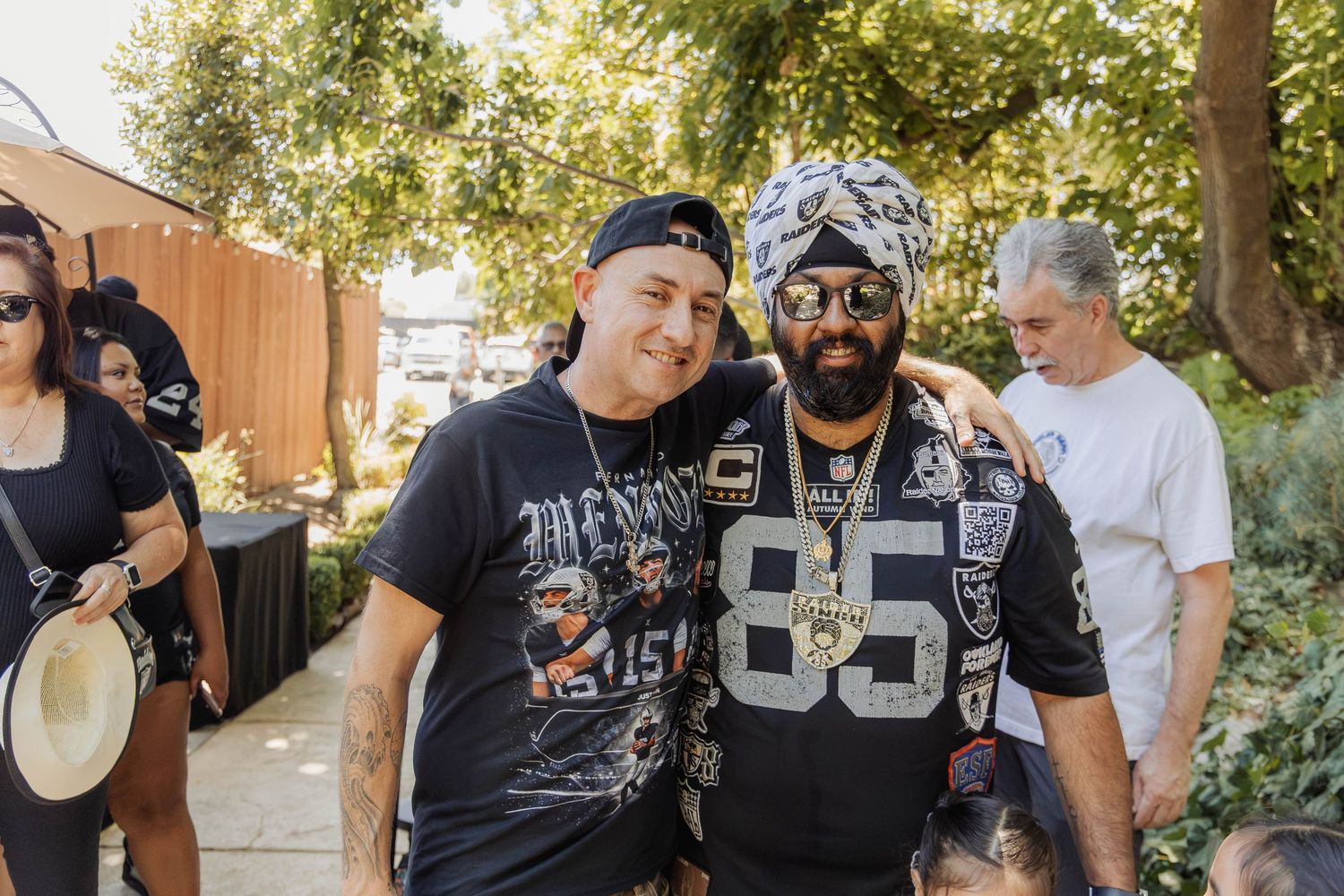 Two men in Raiders gear posing together outdoors at a casual gathering on a sunny day.