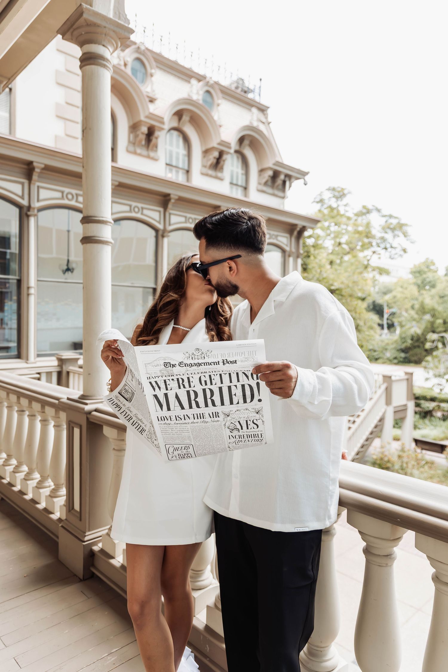 Couple shares intimate moment while holding newspaper announcing their marriage on elegant porch with columns.