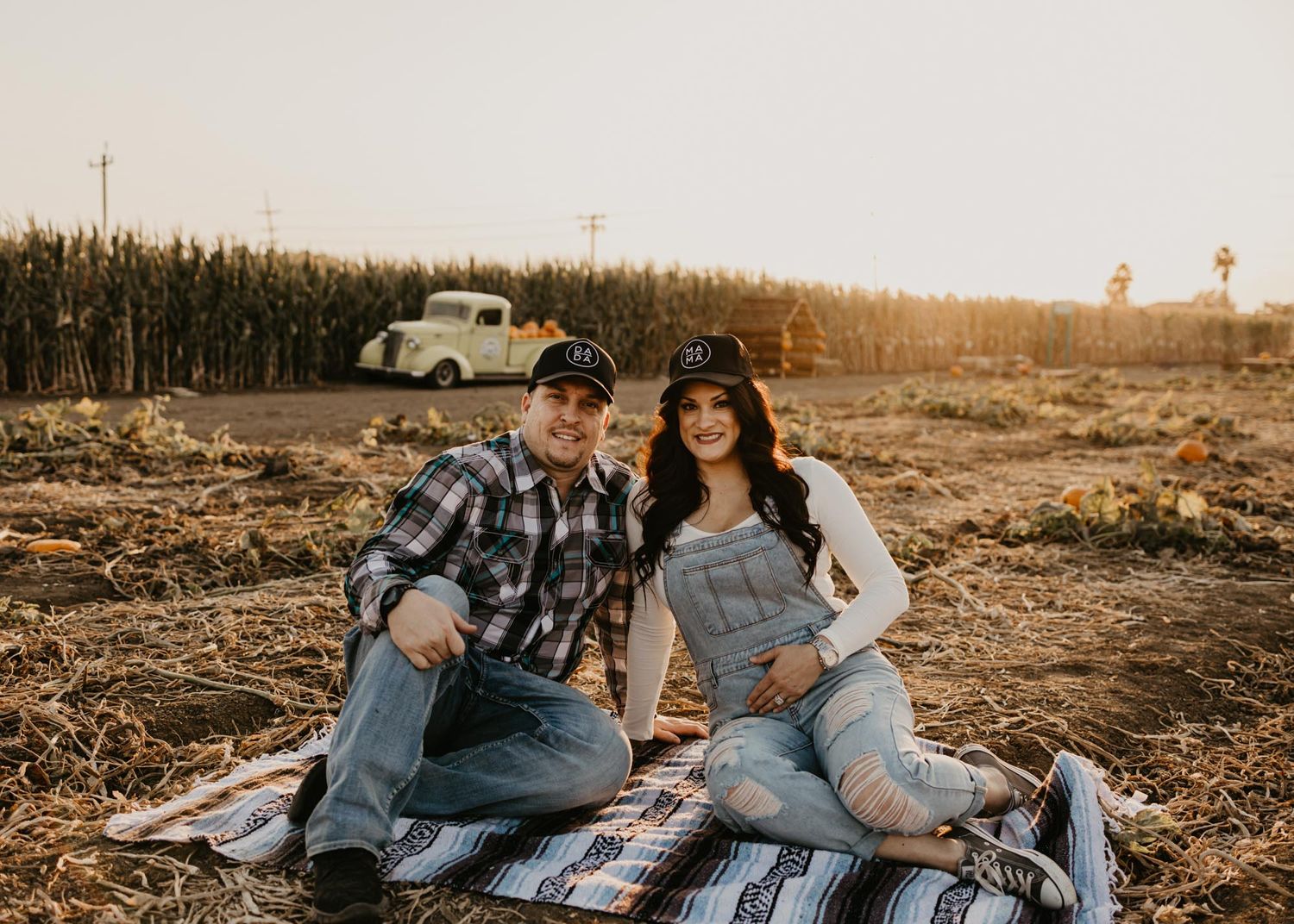 Couple sitting on blanket in field with vintage car and cornfield in background.