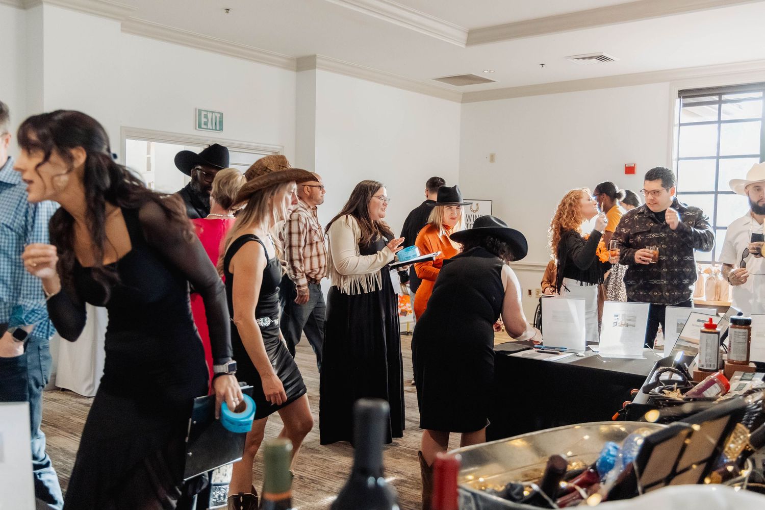 Guests mingle at an indoor wine tasting event, browsing bottles on tables in a bright, modern venue.