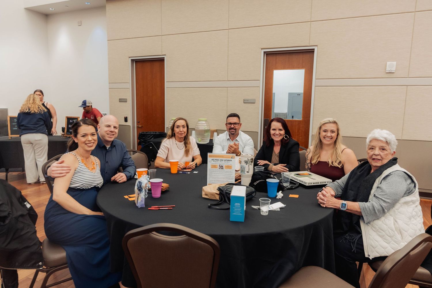 Group of seven adults smiling at a round table during a formal banquet event in a conference hall.