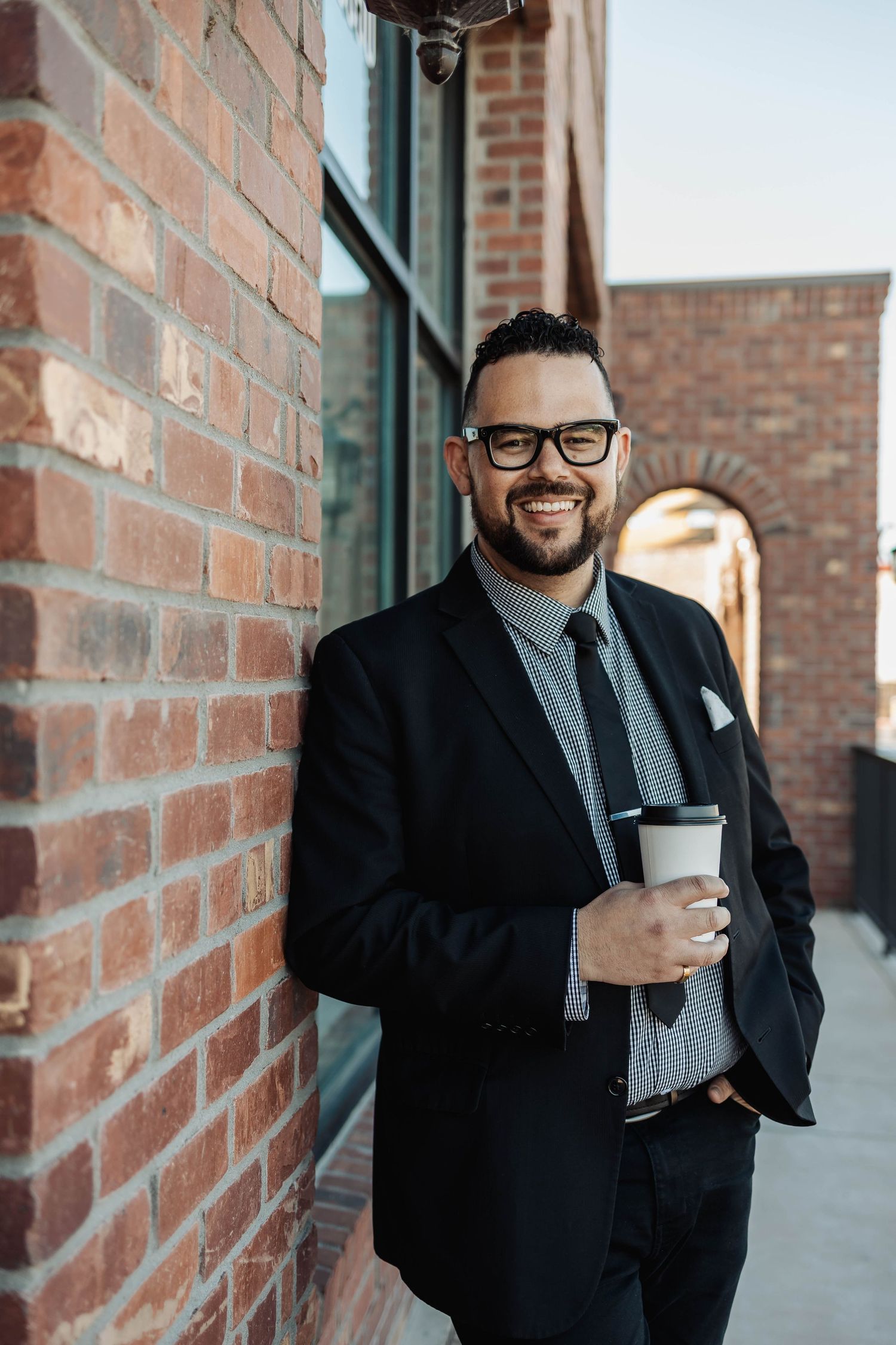 Professional in black suit and glasses holding coffee cup against brick wall.