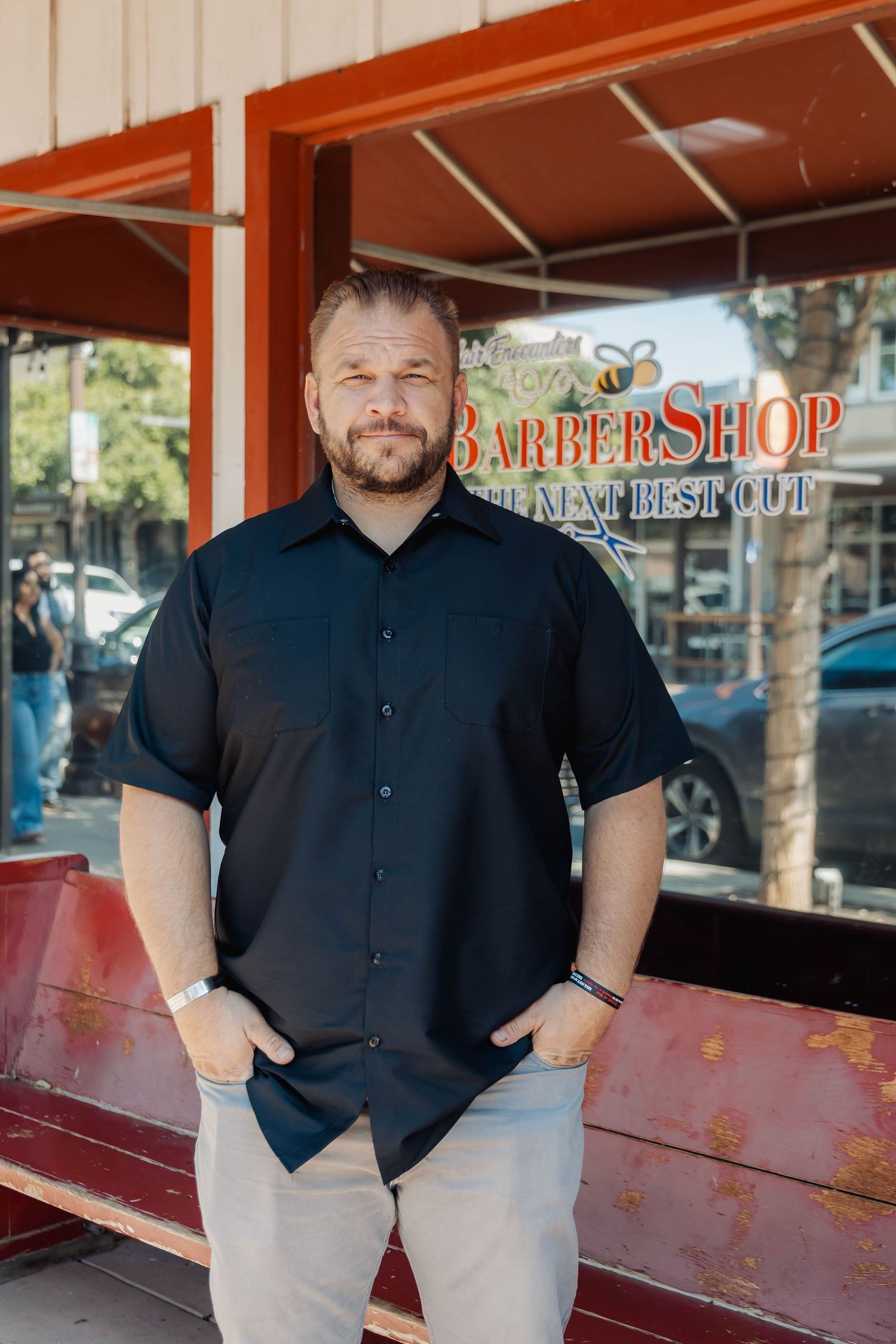 Professional portrait of someone in dark shirt and light pants standing outside barber shop entrance.