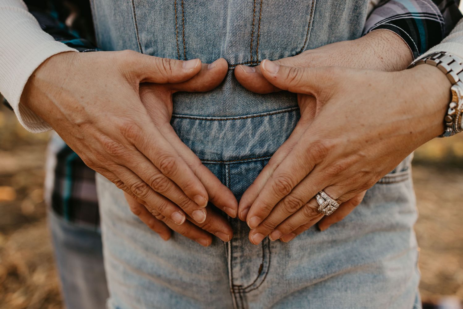 Close-up of hands forming a heart shape on denim overalls with an engagement ring visible.