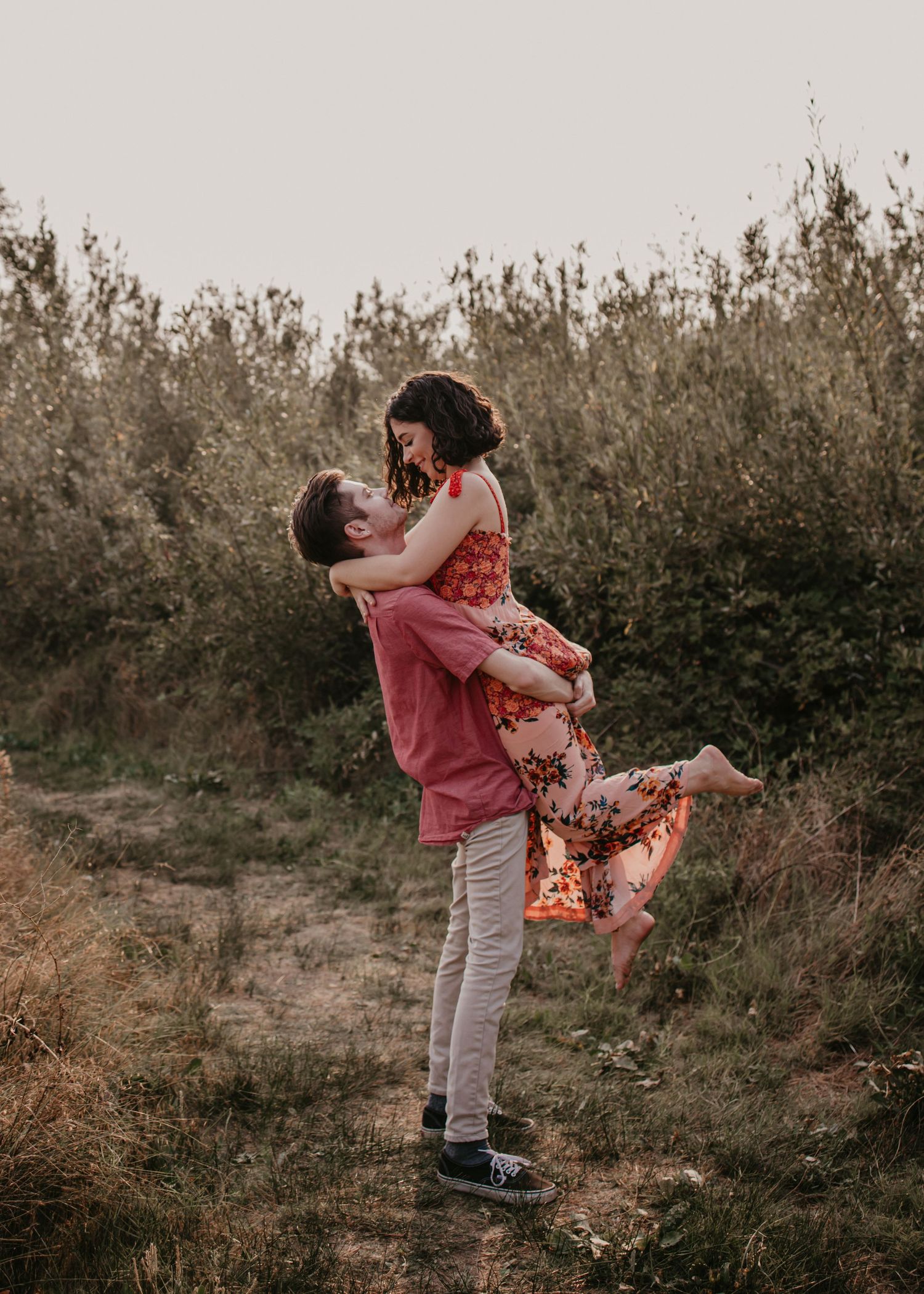 A couple in a floral dress and pink shirt embrace playfully in a natural outdoor setting during sunset.