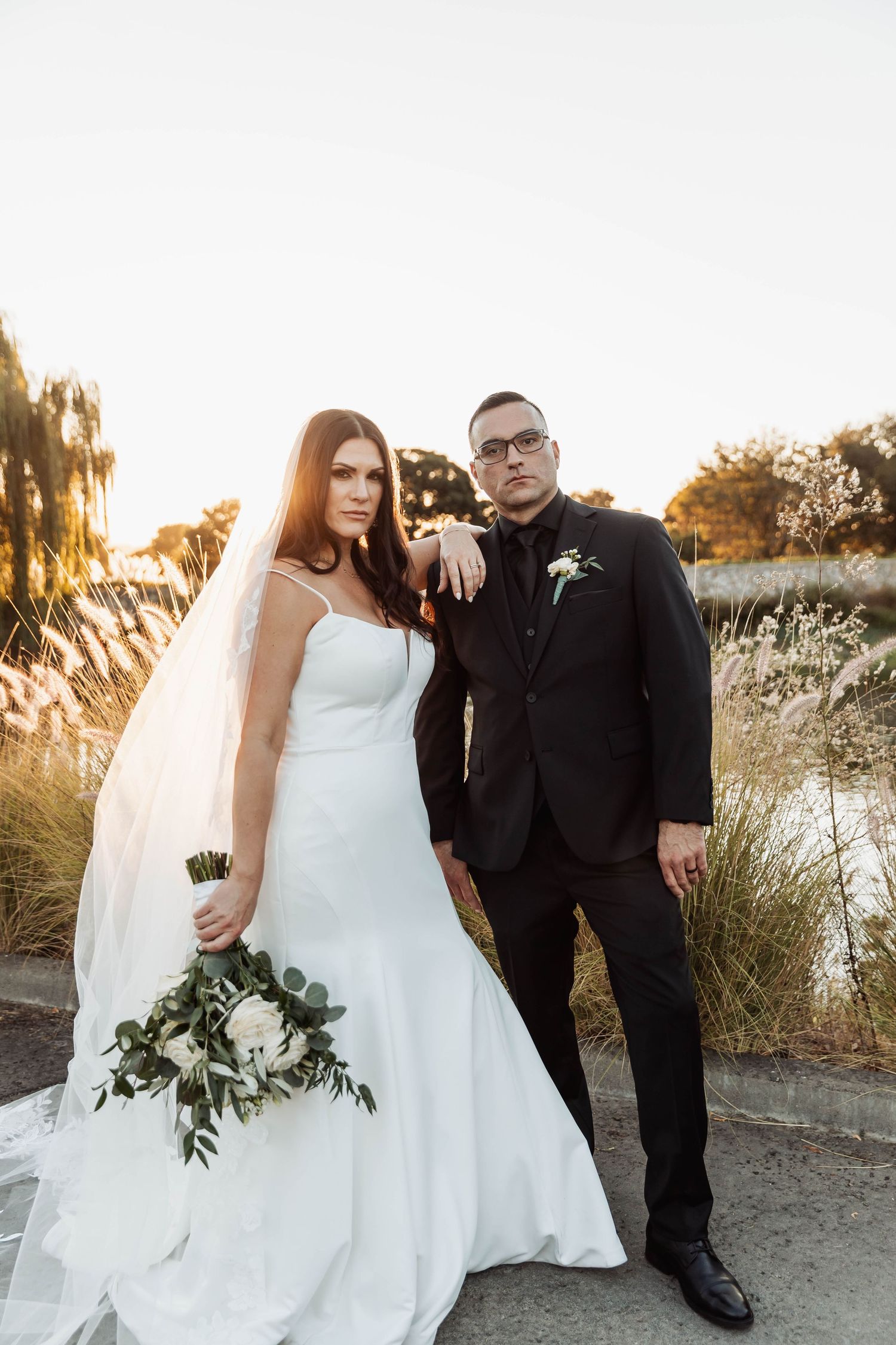 Newlyweds pose together against sunset backdrop holding greenery bouquet.