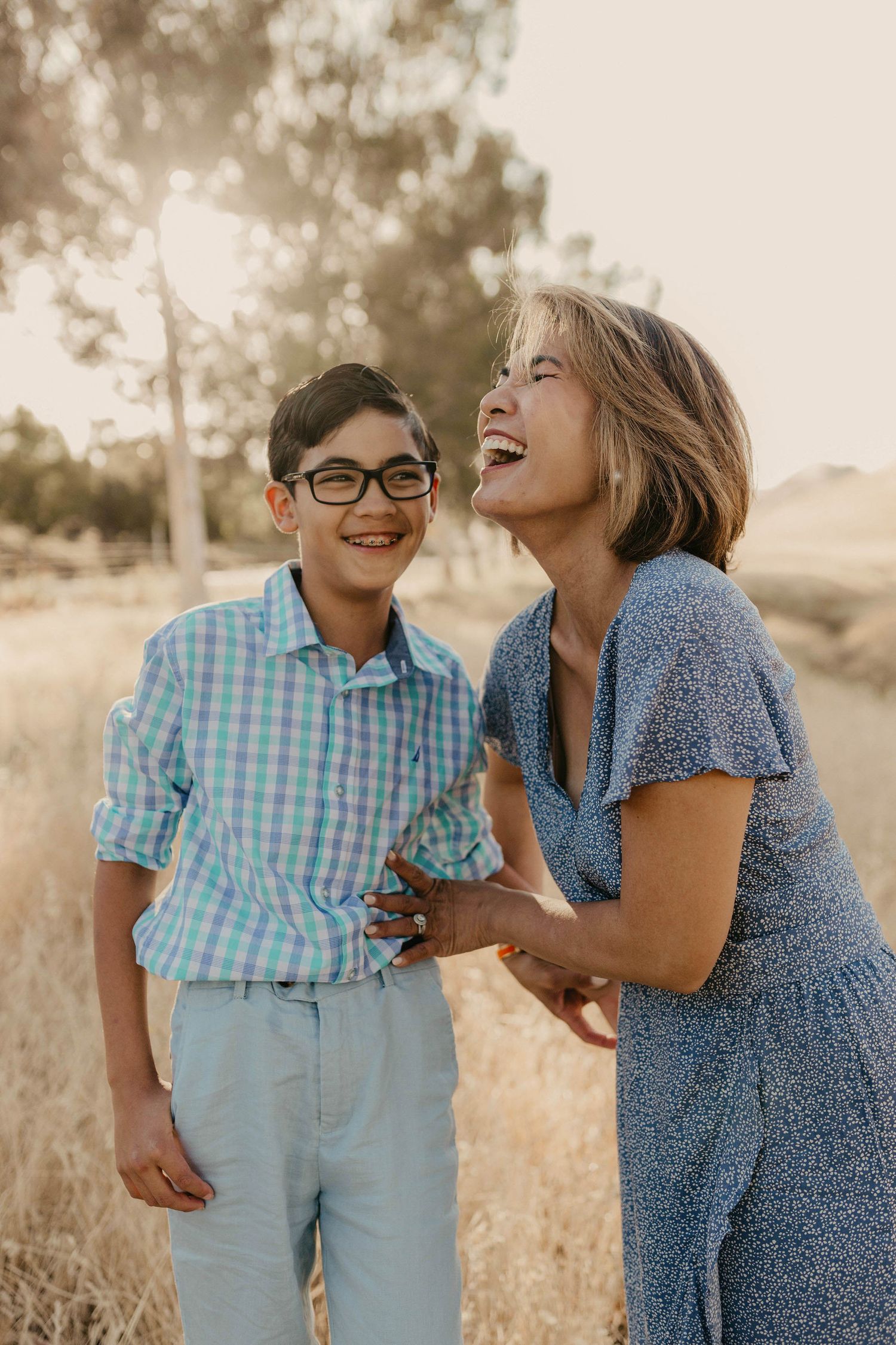 A woman and young boy with glasses laugh together outdoors in a golden field during a warm family photo session.
