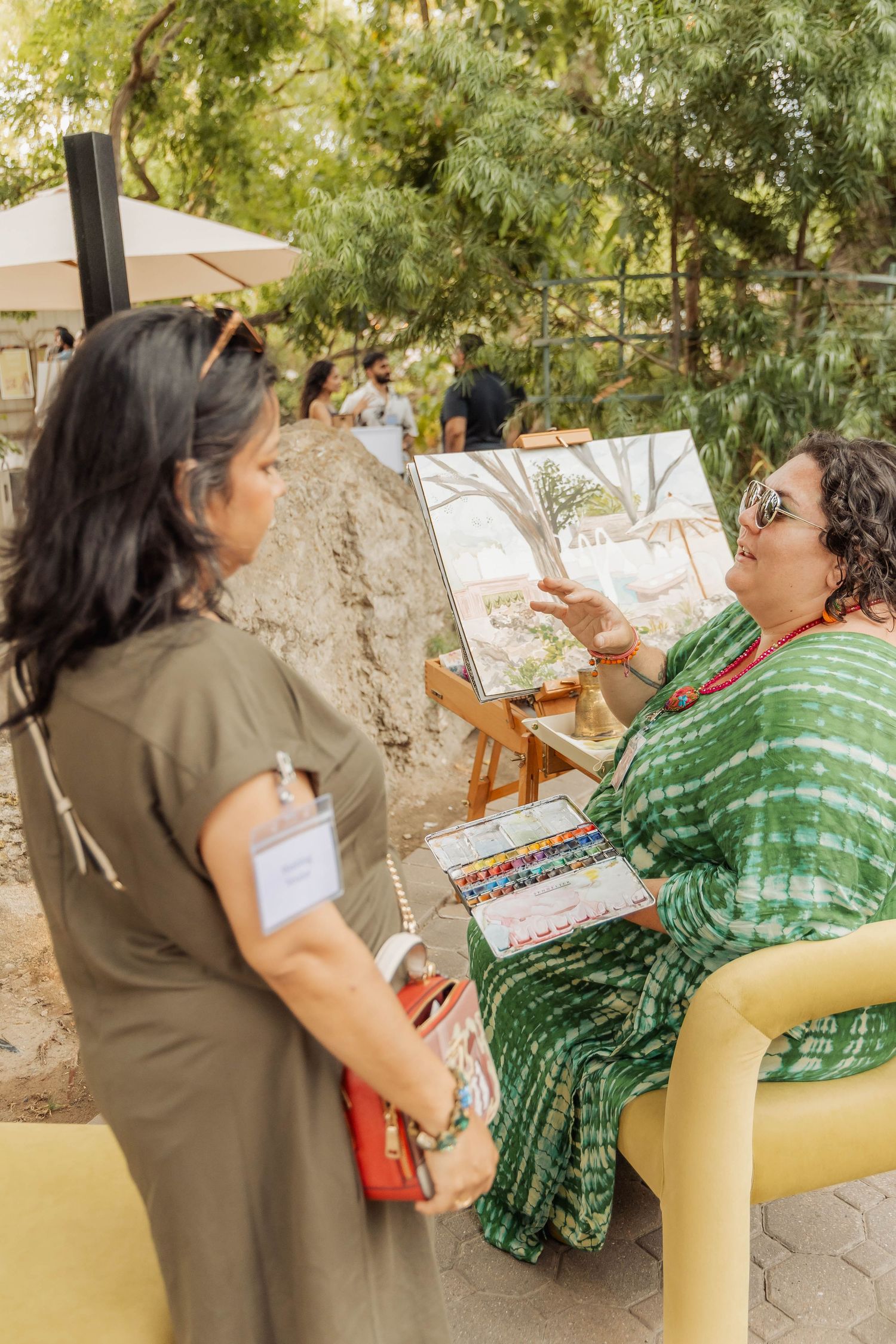 Artist in green sari discusses watercolor painting on easel with woman in olive dress at outdoor art event.