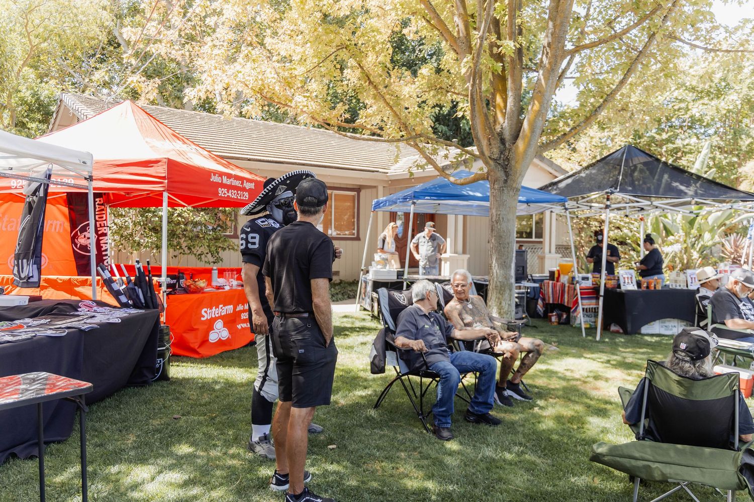 Outdoor fan event with colorful vendor tents, attendees relaxing on chairs under shady trees on a sunny day.