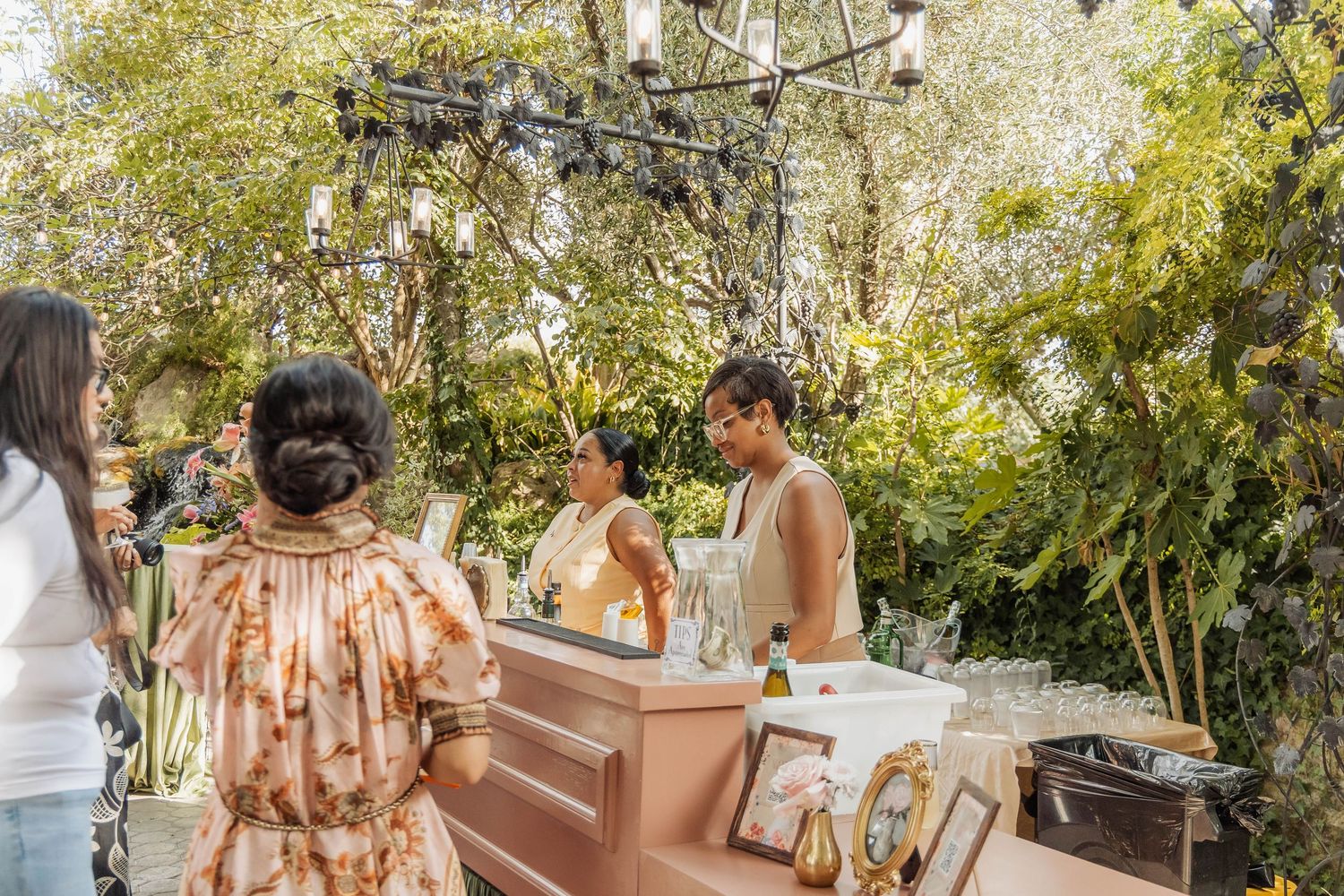 Women gather at an outdoor bar surrounded by lush greenery and hanging chandeliers at an elegant garden event.