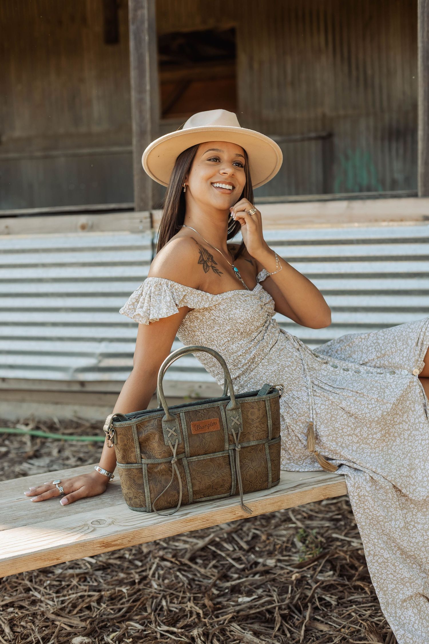 White floral dress paired with tan hat and vintage leather bag in rustic setting.