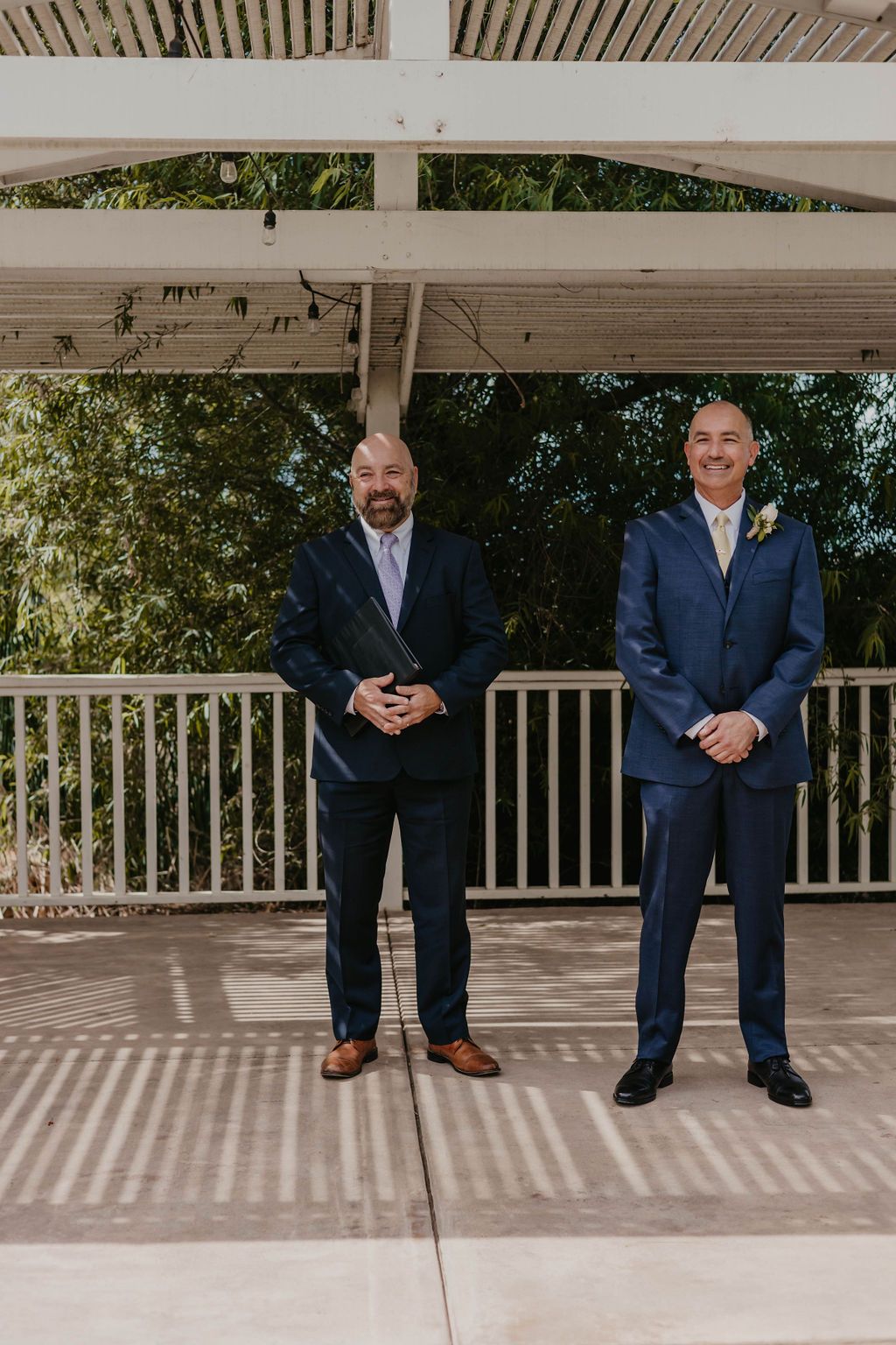 Two people in suits stand on a covered patio during a wedding ceremony.