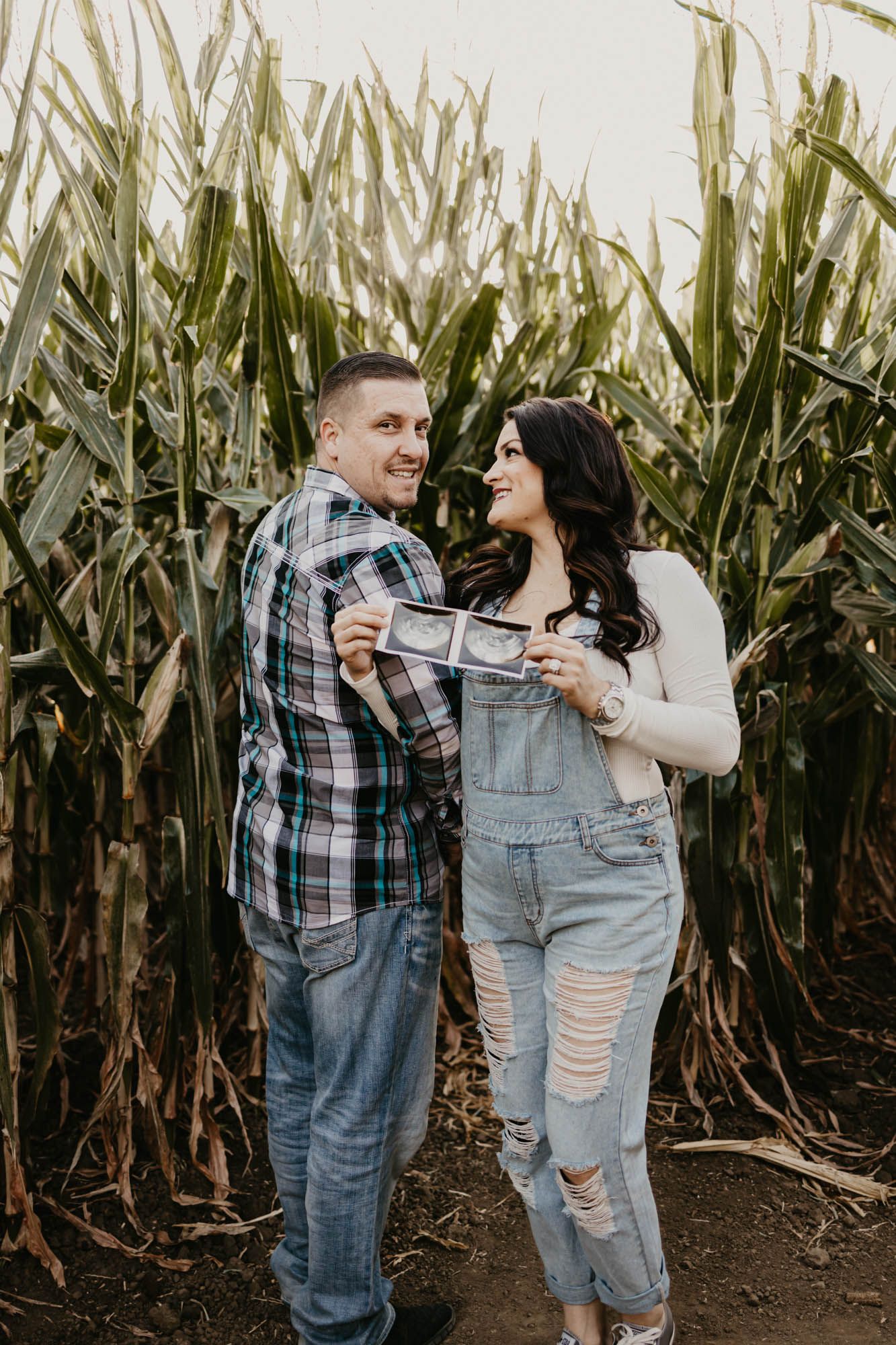 Happy couple laughing together while displaying their pregnancy announcement photos in a corn maze.