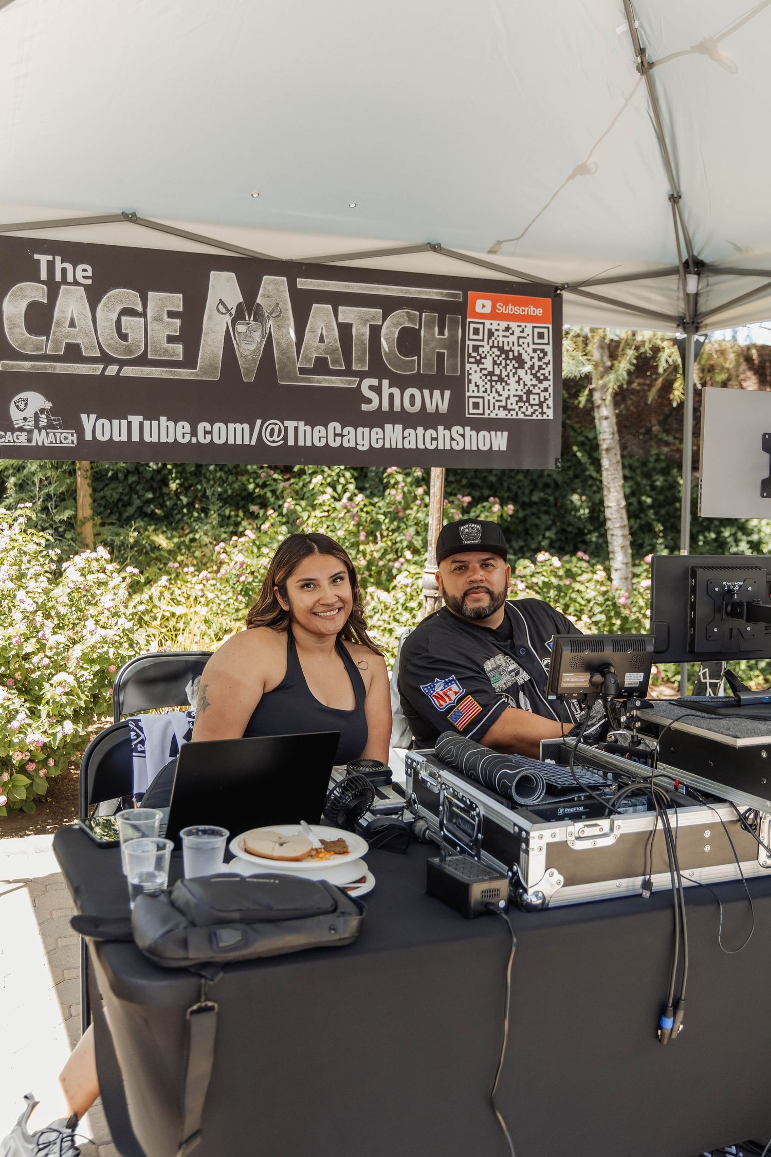 Two smiling hosts at The Cage Match Show booth with DJ equipment and laptop at an outdoor event.